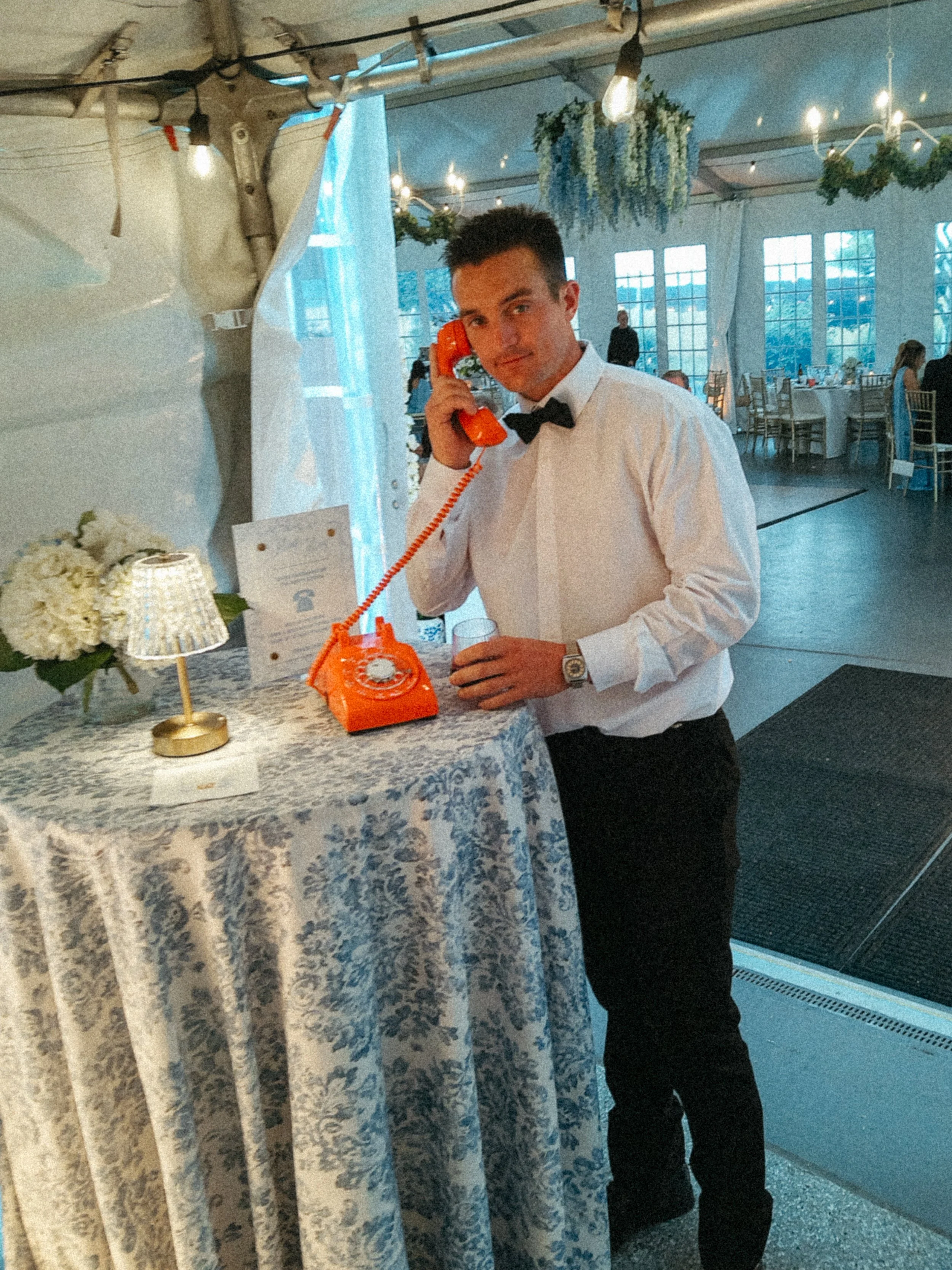 A young man dressed in a white shirt with a black bow tie, holding an orange vintage rotary phone, standing next to a round table with a white and blue floral tablecloth, in a decorated indoor event venue with tables, chairs, hanging plants, and string lights.