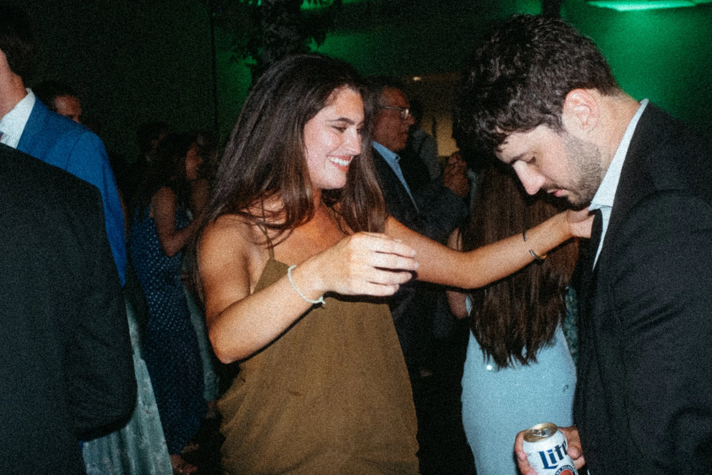 People socializing at a party, two women in the center are smiling and chatting, one holding a beer can. Others are in the background, some wearing suits and dresses. Taken on a disposable camera to give a nostalgic, vintage look.