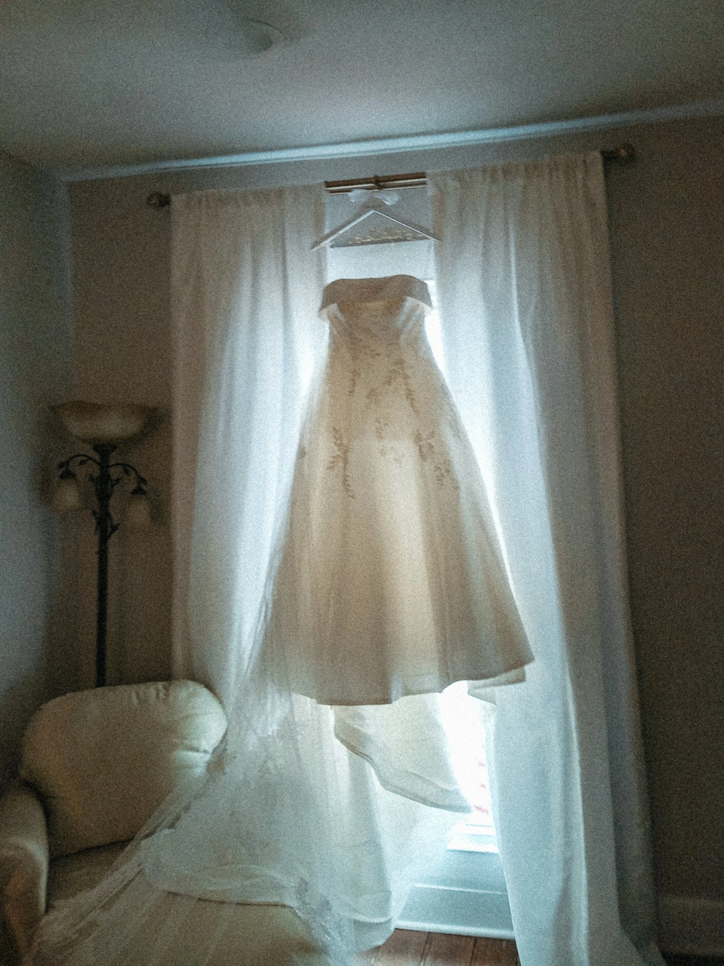 A wedding dress hangs on a hanger in front of a window with light-colored curtains in a cozy room. A sofa and a lamp are partially visible in the room. Taken on a disposable camera to give a nostalgic, vintage look.