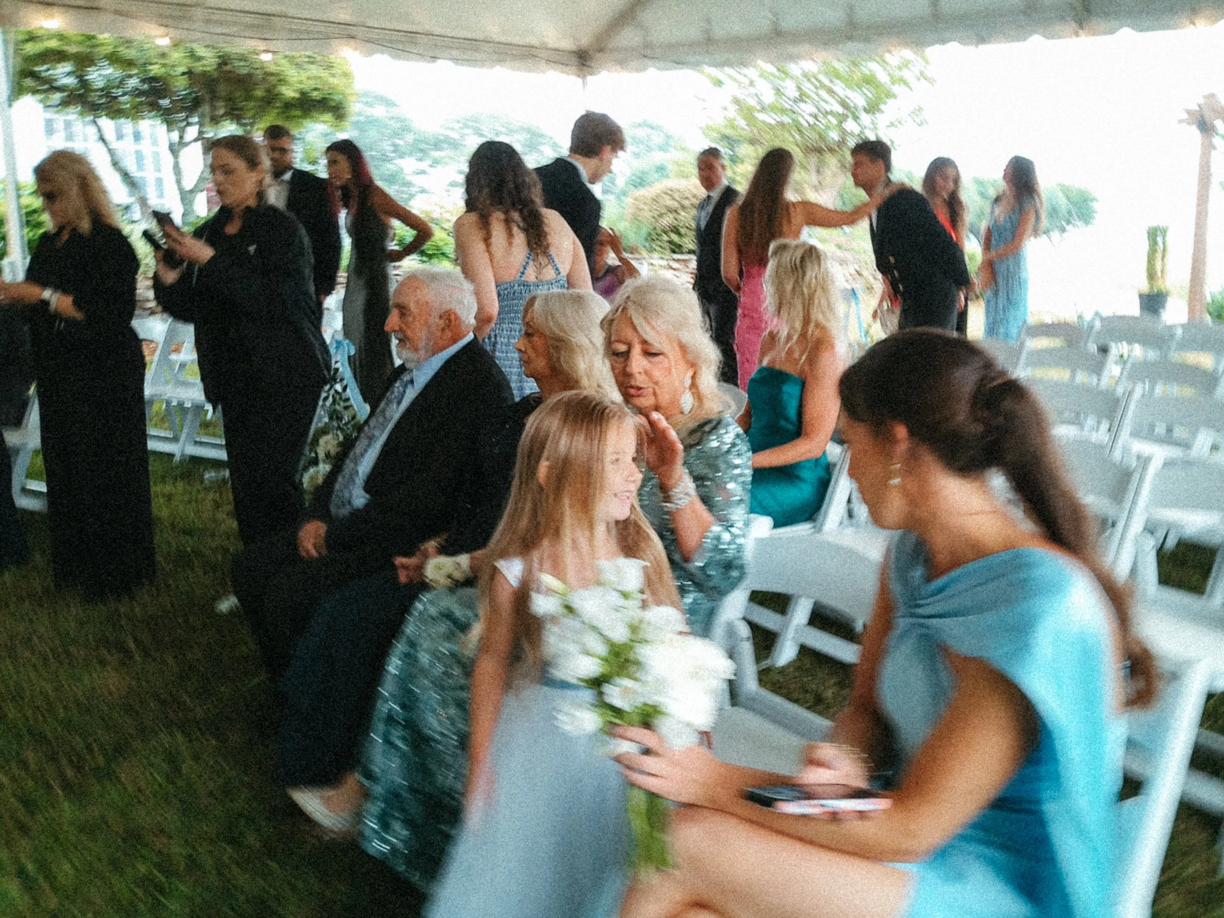 People at a social gathering under a tent, some standing and some sitting, with greenery and trees in the background. Taken on a disposable camera to give a nostalgic, vintage look.