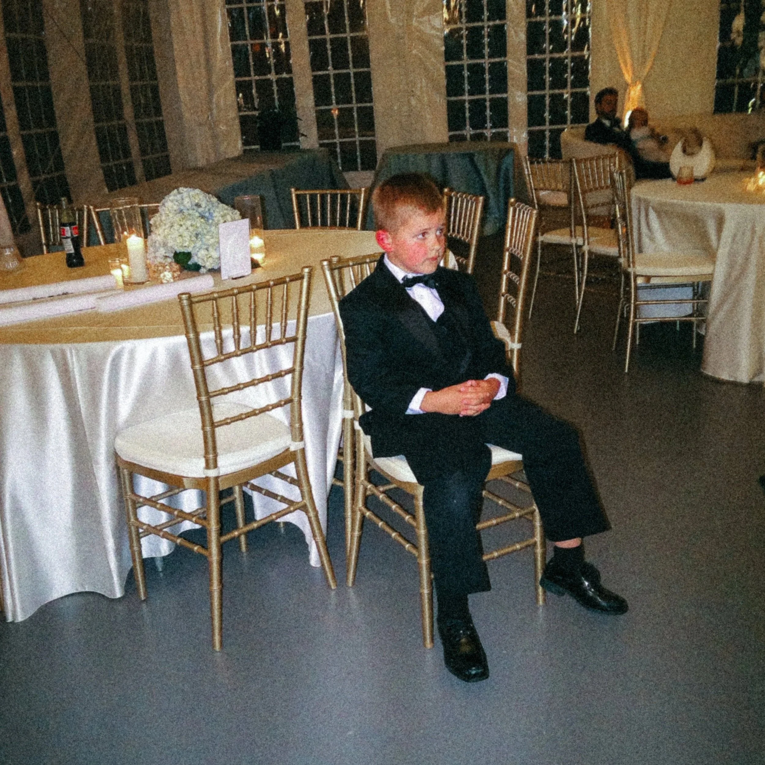 A young boy dressed in a tuxedo sitting at a banquet table with a floral centerpiece, candles, and menu cards in a dimly lit room with large windows and curtain drapes, with a man and woman sitting at another table in the background.