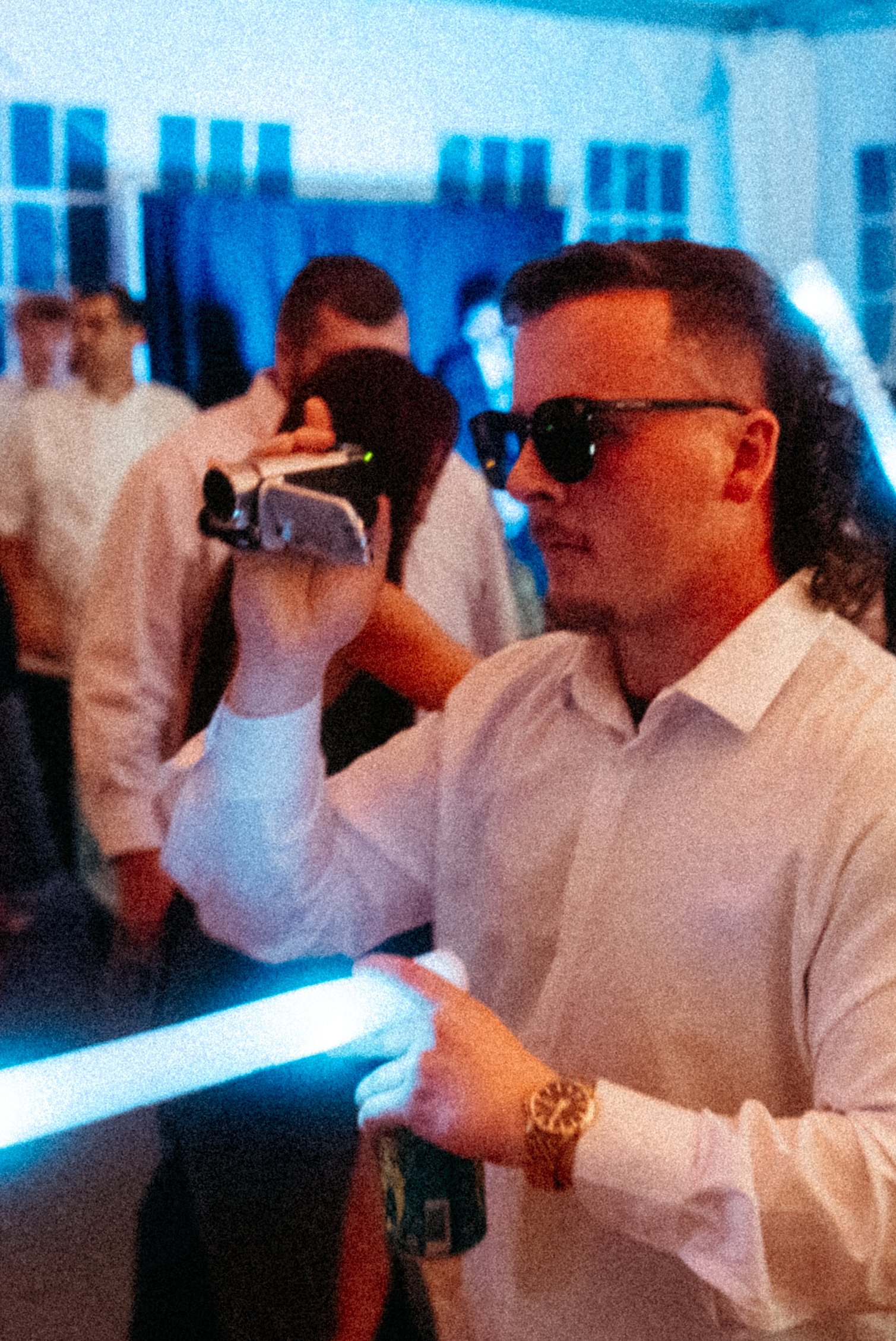 A man in white shirt and sunglasses holding a camera capturing a moment at a social gathering with people in the background in a dimly lit room with blue lighting. Taken on a disposable camera to give a nostalgic, vintage look.
