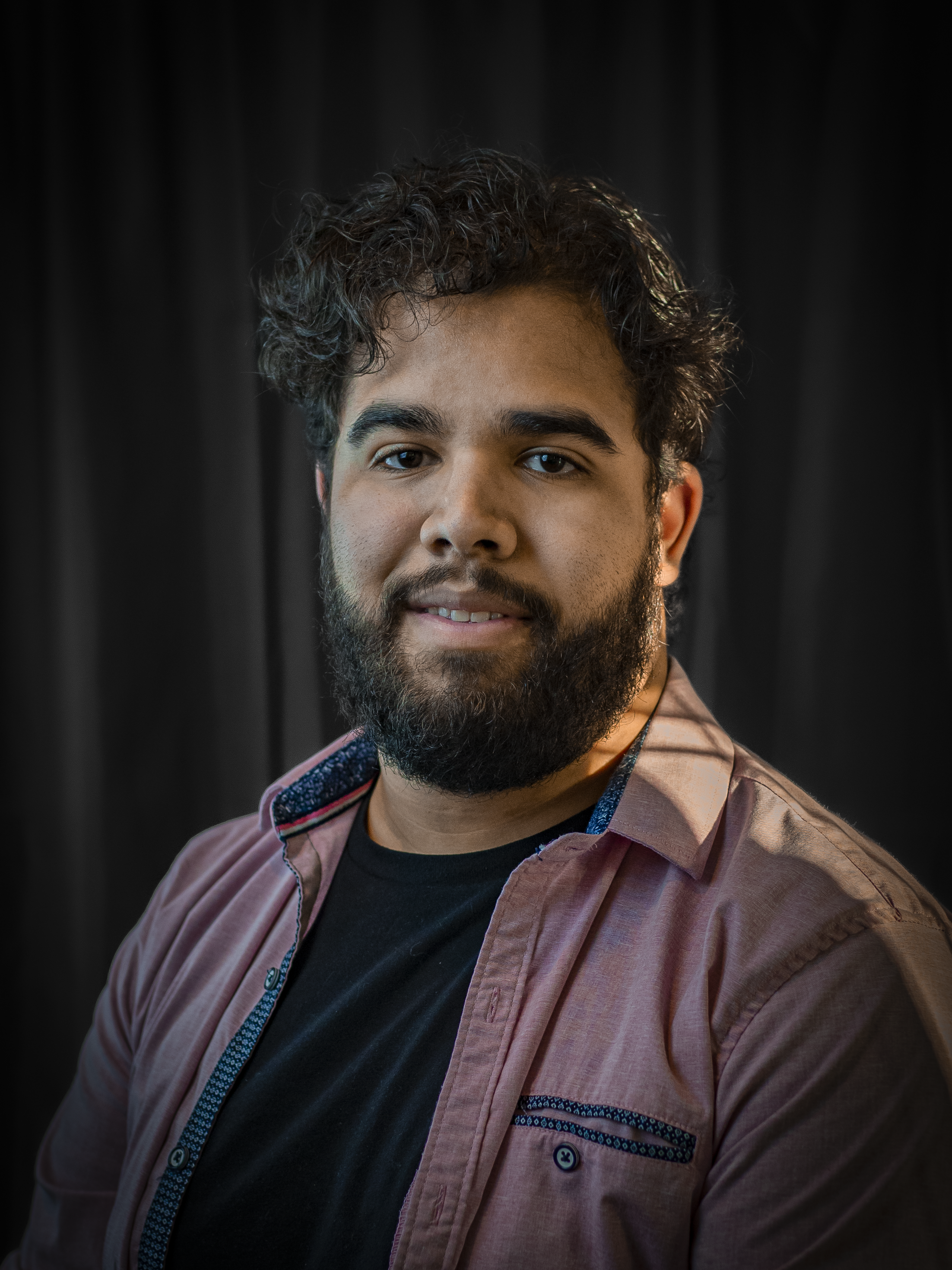 Portrait of a man with curly dark hair, beard, and mustache, smiling slightly, wearing a black t-shirt and a pink button-up shirt with decorative blue patterns on the inside collar and pockets, in front of dark curtains.
