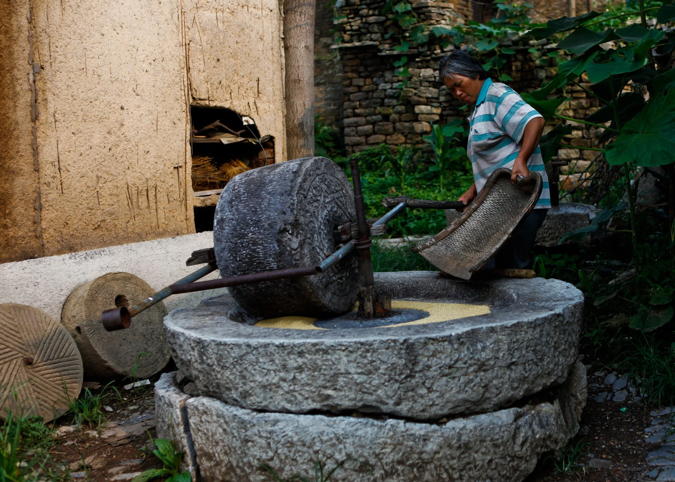 A woman operating a stone mill grinder outdoors, surrounded by lush green plants and traditional stone buildings.