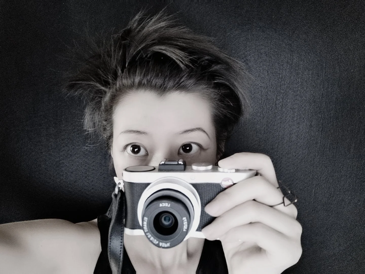A woman with short, tousled hair holding a camera in front of her face, capturing a photo. She is looking directly at the camera, with a dark background.