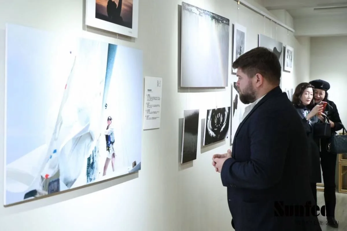 A man with a beard and dark hair, dressed in a black suit, is observing artwork on a gallery wall. In the background, two women are looking at their phones; one is wearing a black beret and the other has long dark hair.