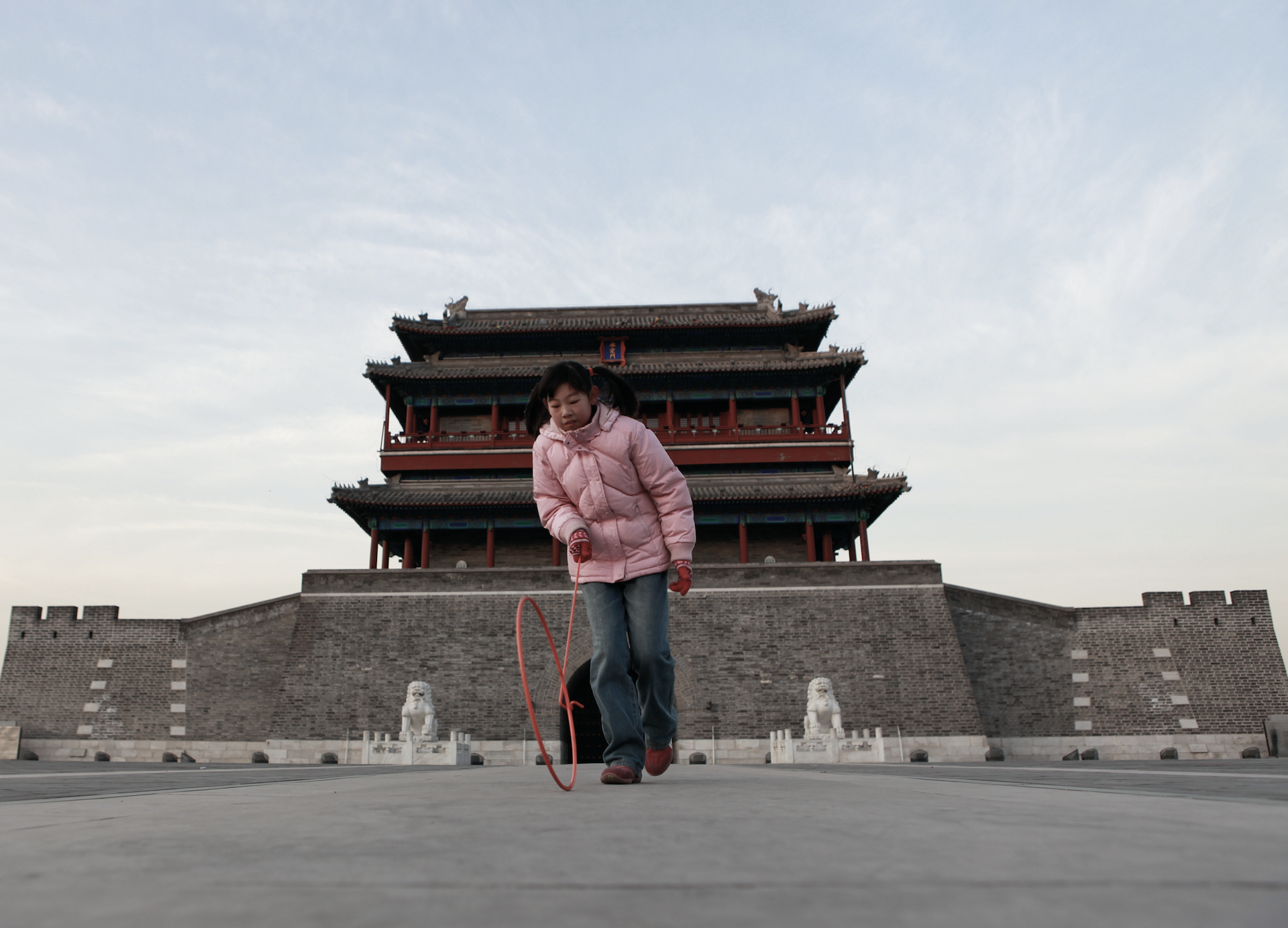 A young girl in a pink jacket and blue jeans skipping rope in front of an ancient Chinese building with red wooden structure and gray stone walls under a cloudy sky.