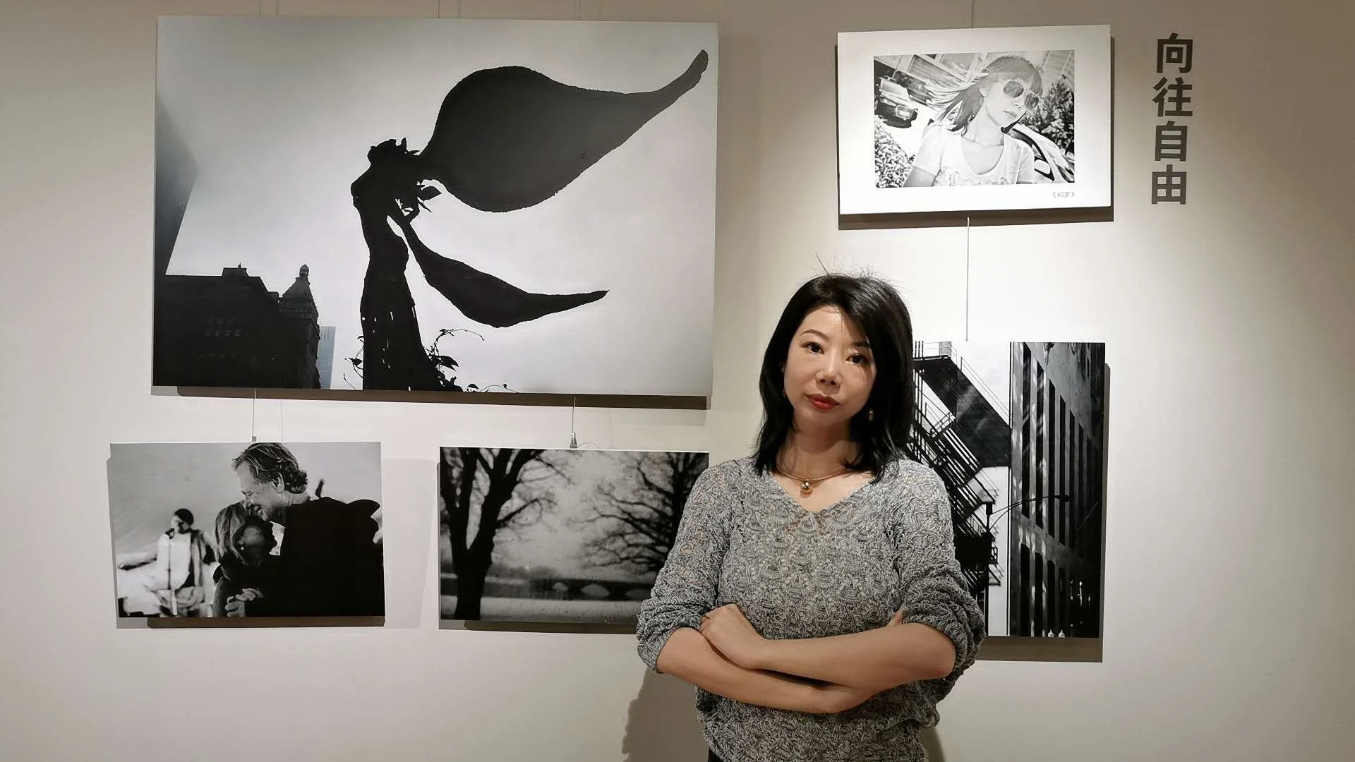 A woman with black hair wearing a patterned long-sleeve top standing with her arms crossed in front of a wall with black and white photographs and artwork, including a silhouette of a woman with flowing hair, a woman with sunglasses, and cityscape images, in an art gallery.