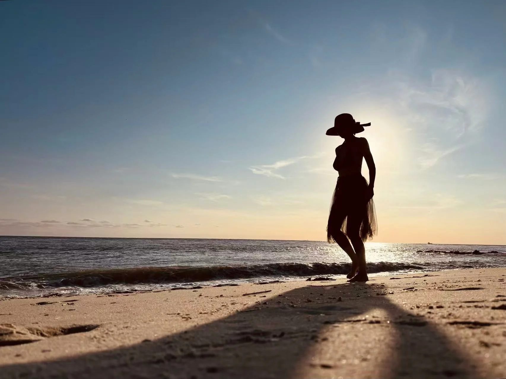Cinematic coastal lifestyle portrait of a woman in a sun hat and flowing skirt walking on the beach at golden hour, ocean waves and warm light creating a serene atmosphere, photography by Jing Xiao