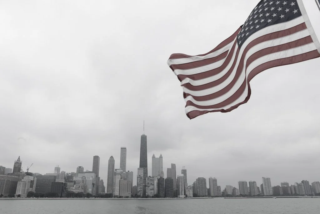 American flag waving in front of a city skyline with tall buildings and a cloudy sky.