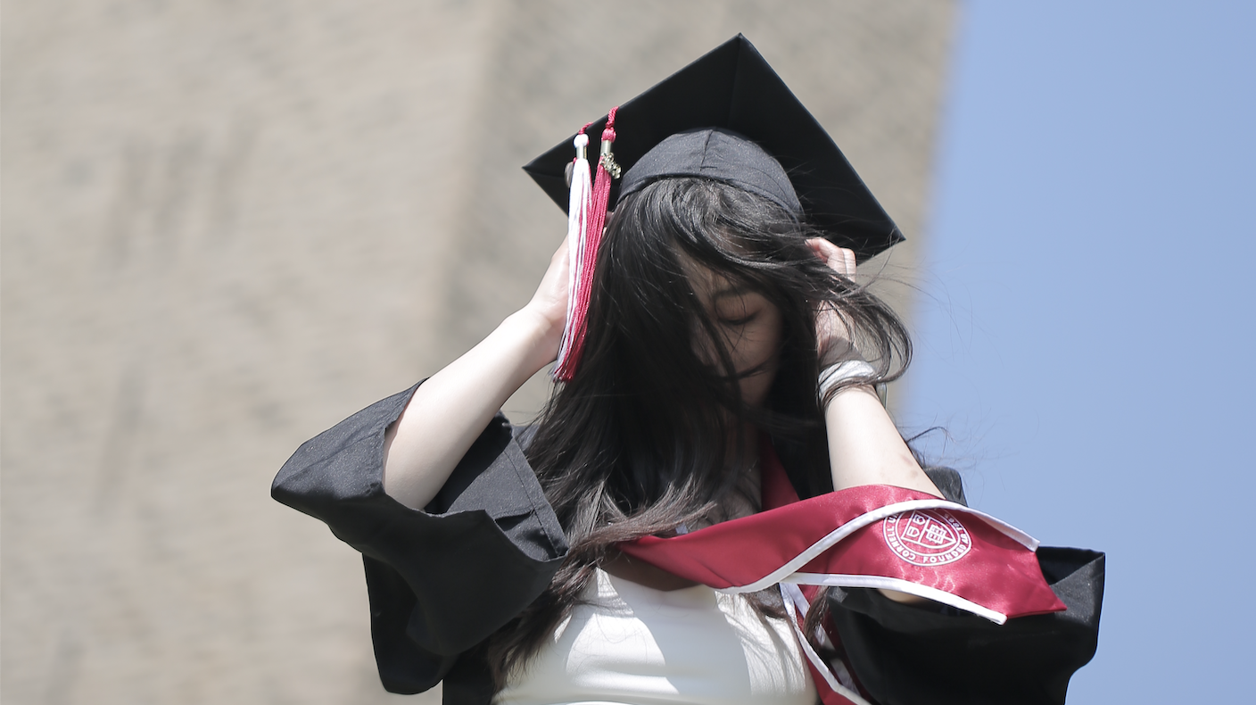 Cinematic graduation portrait of a young woman adjusting her cap outdoors in natural light, wearing a cap and gown with soft blurred background and blue sky, photography by Jing Xiao