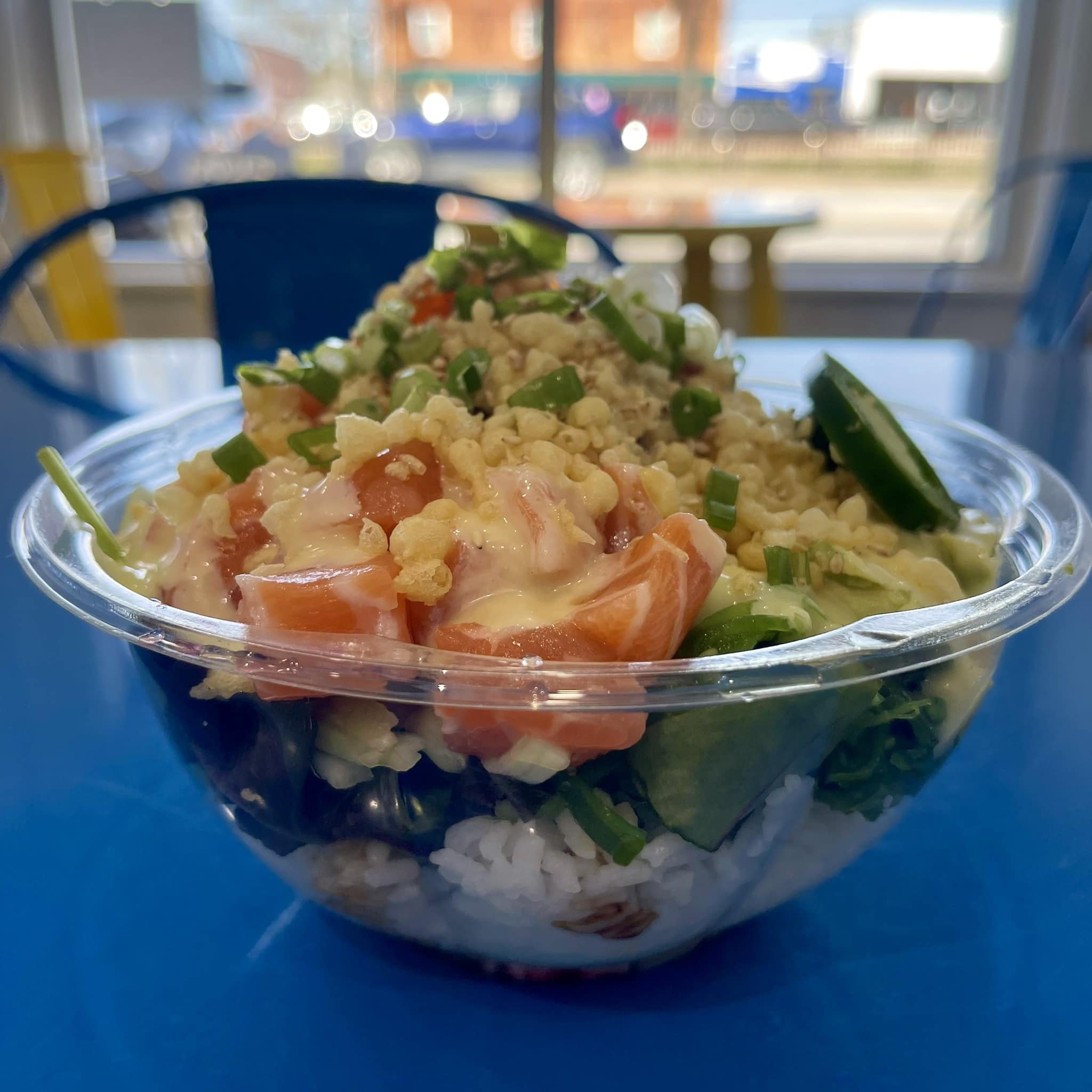 Close-up of a bowl of couscous salad with chopped green onions, diced tomatoes, and what appears to be shredded cheese, set on a blue table with a blurred view of a window and streets outside in the background.