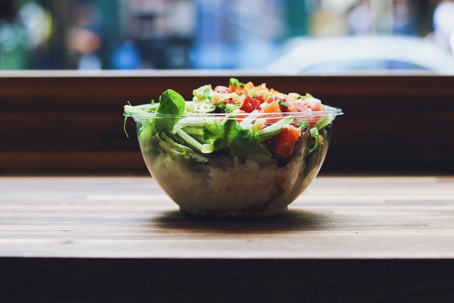 Vegetable salad in a clear plastic bowl on a wooden surface with a window in the background