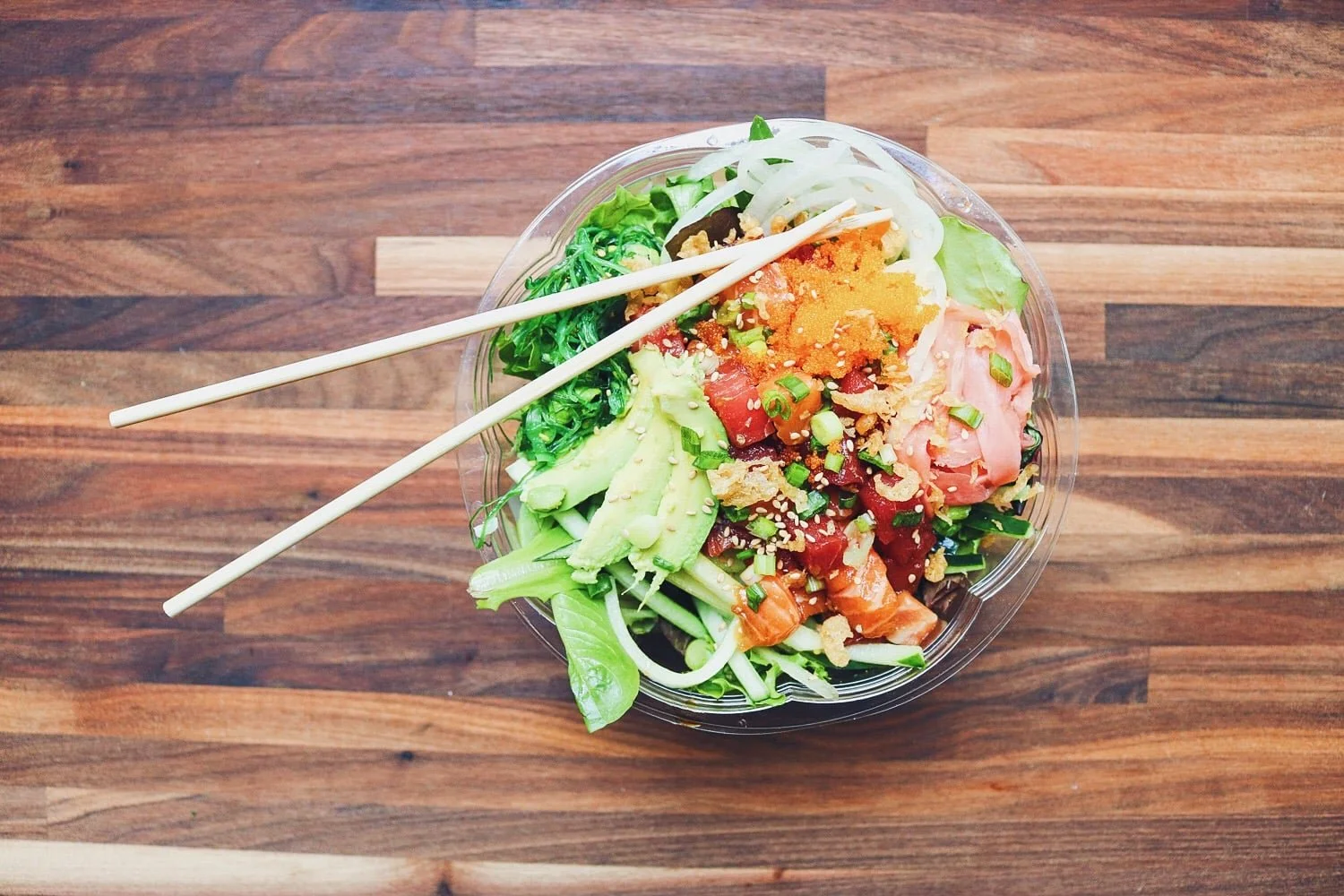 A bowl of sushi salad with fresh vegetables, sliced avocado, chopped onions, and chopped raw fish topped with sesame seeds, with a pair of chopsticks resting on top, on a wooden table.