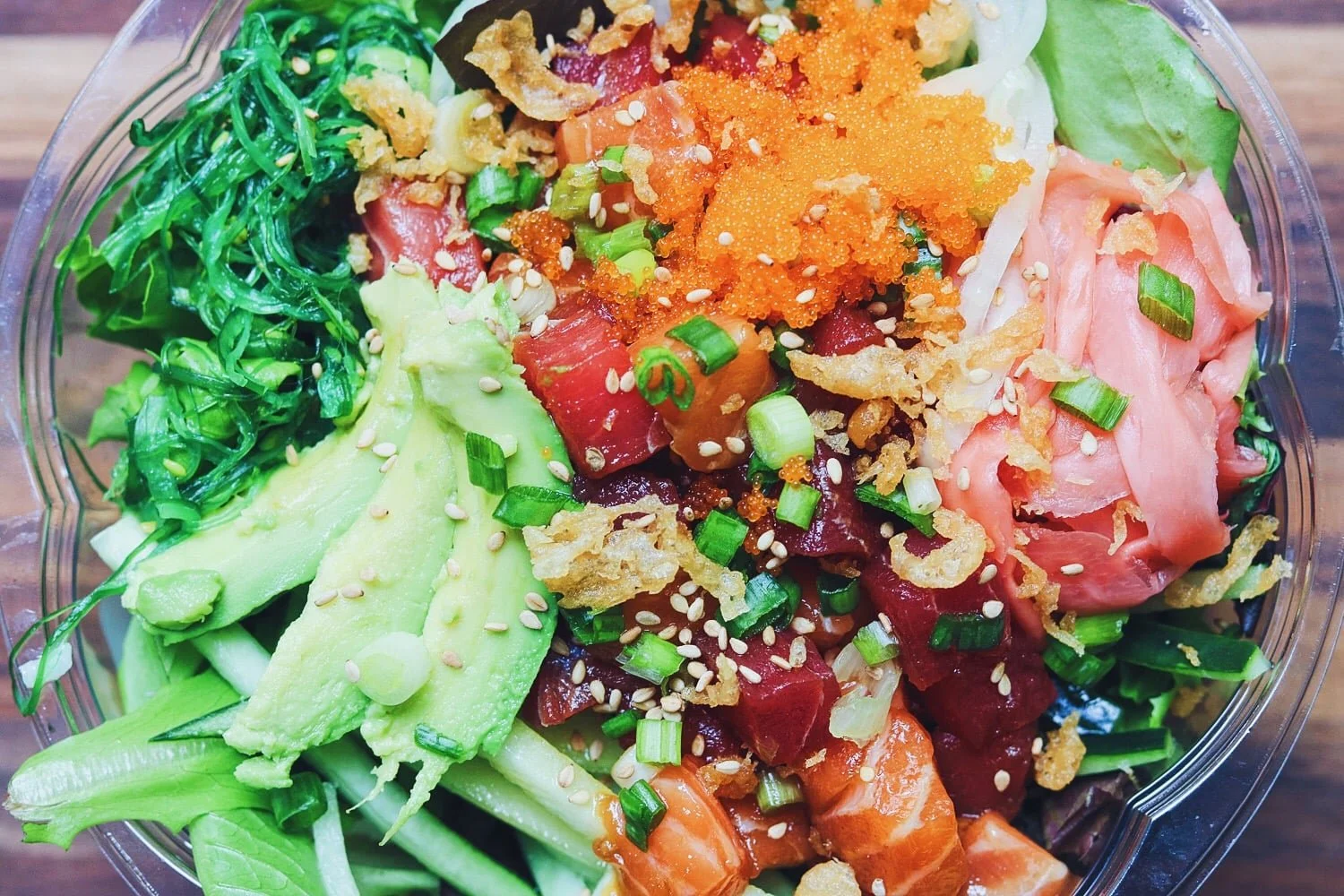 A close-up of a bowl of poke salad with diced raw salmon, tuna, avocado slices, chopped green onions, seaweed, fish roe, topped with sesame seeds and crispy garlic bits.