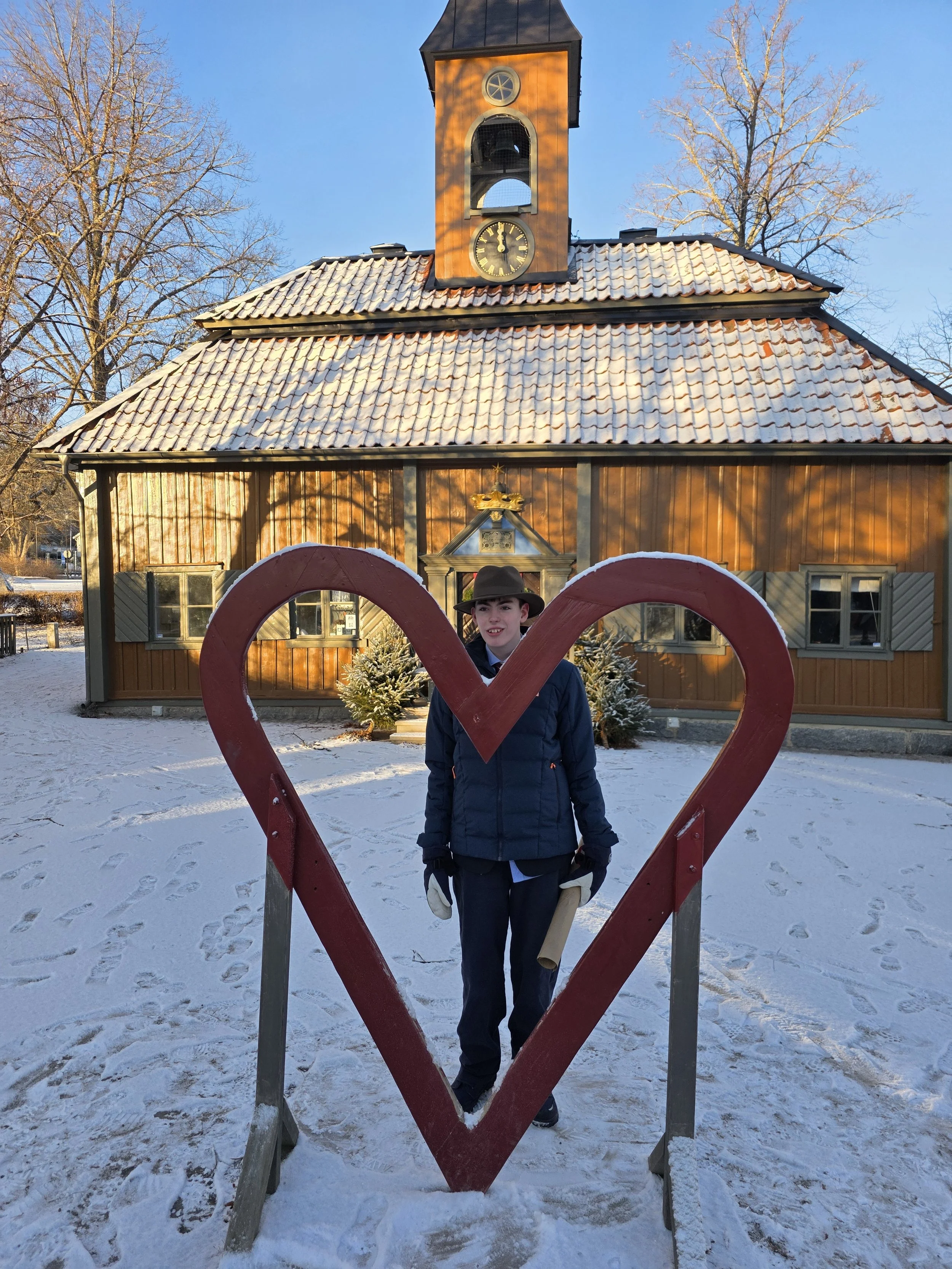 A person dressed in a dark coat, gloves, and a hat standing in front of a large, red heart-shaped frame outdoors in a snowy area. Behind them is a wooden building with a clock tower, decorated with snow, and leafless trees are visible against a clear blue sky.