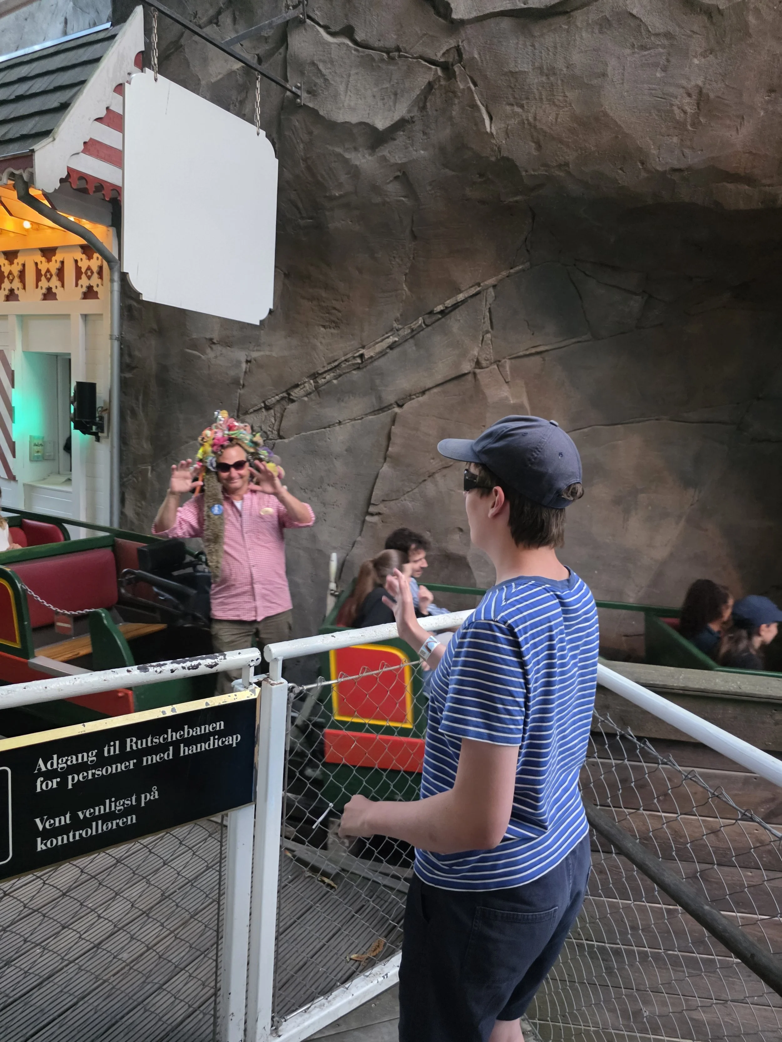 A woman with a long braided hair hair decorated with colorful flowers, wearing sunglasses and a pink shirt, smiling and waving. A boy in a striped blue and white shirt and a cap is standing nearby, looking at her. They are at an amusement park, possibly near a roller coaster or ride, with a rocky wall in the background and a sign with Norwegian text about accessibility.