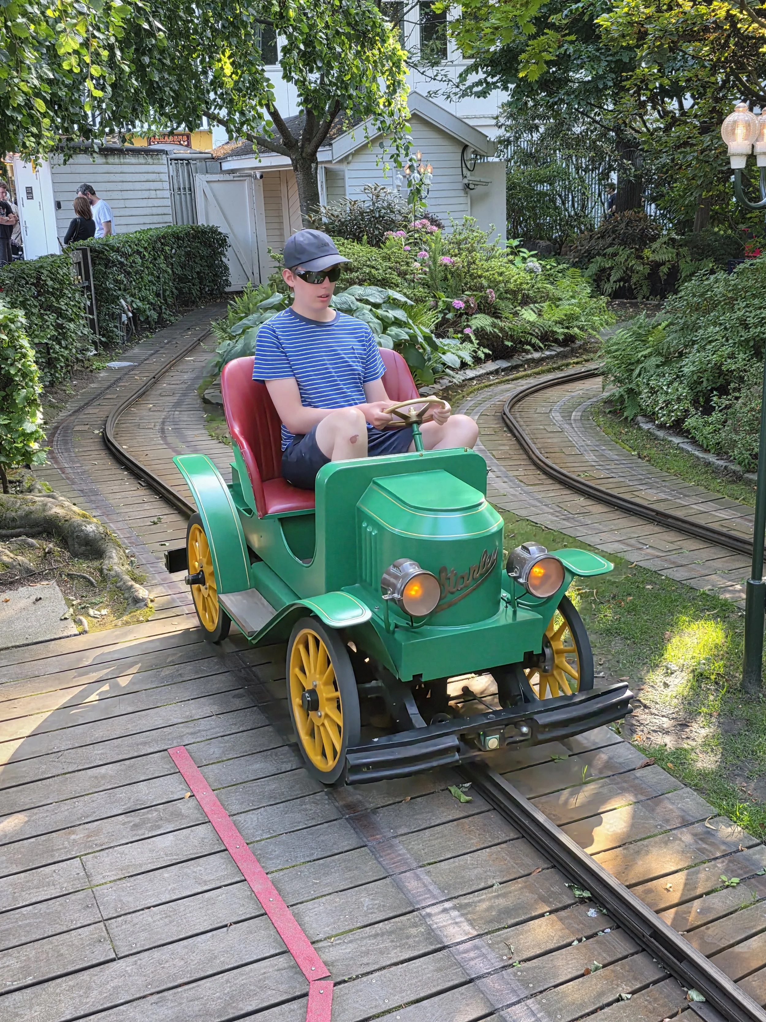 A boy wearing a striped blue t-shirt, black shorts, a baseball cap, and sunglasses is riding a small green and yellow toy train on a track through a garden with lush bushes and trees. In the background, there are a white shed, a hedge, and two people talking.