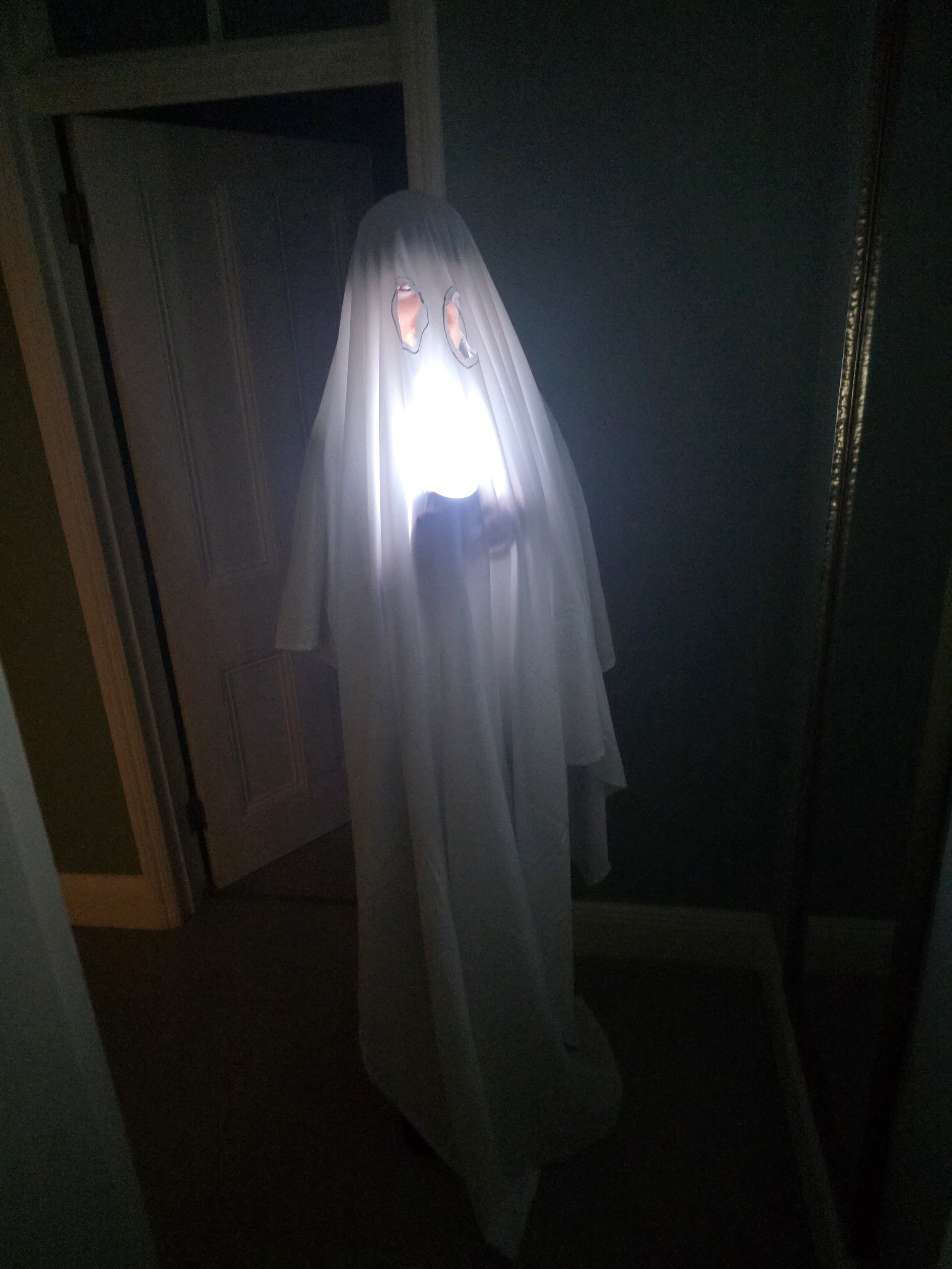 A white ghost decoration with holes for eyes and a mouth, illuminated from below, standing in a dark room.