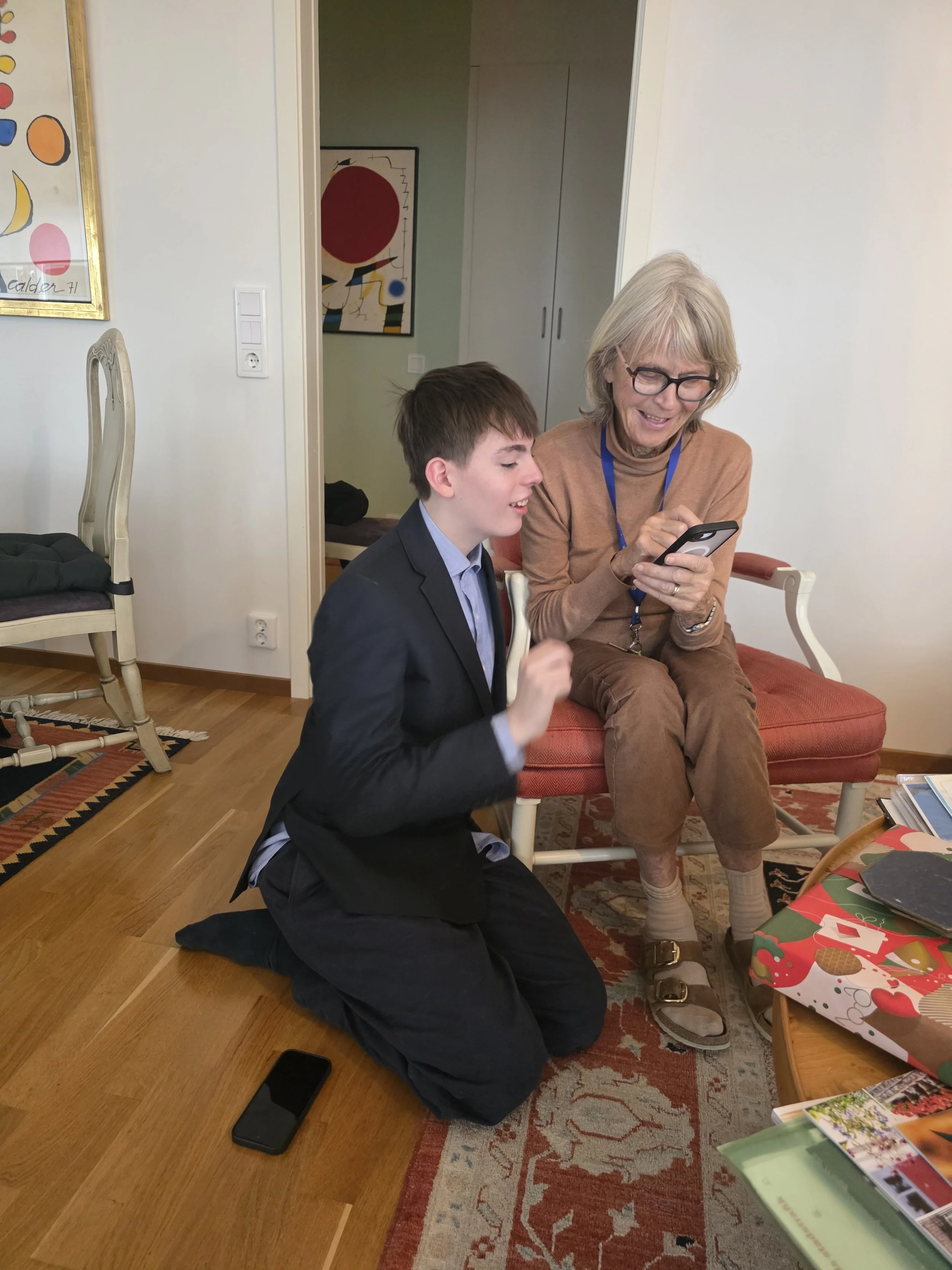 A young man kneeling on the floor with a black shirt and pants, and an older woman sitting on a red cushioned chair, wearing glasses and a brown outfit, looking at a smartphone. They are in a room with wooden flooring, a patterned rug, and a table with various items, including a Christmas-themed gift. There are paintings on the walls and a closet in the background.