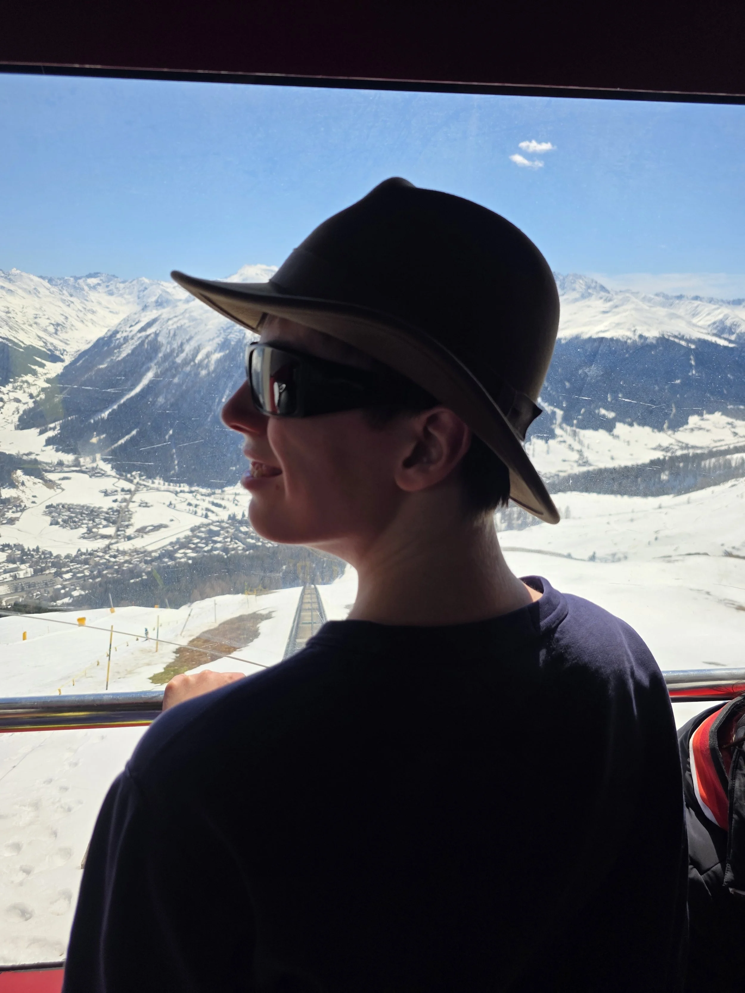 Person wearing a wide-brimmed hat and sunglasses, smiling, with snow-covered mountains and blue sky outside a window.