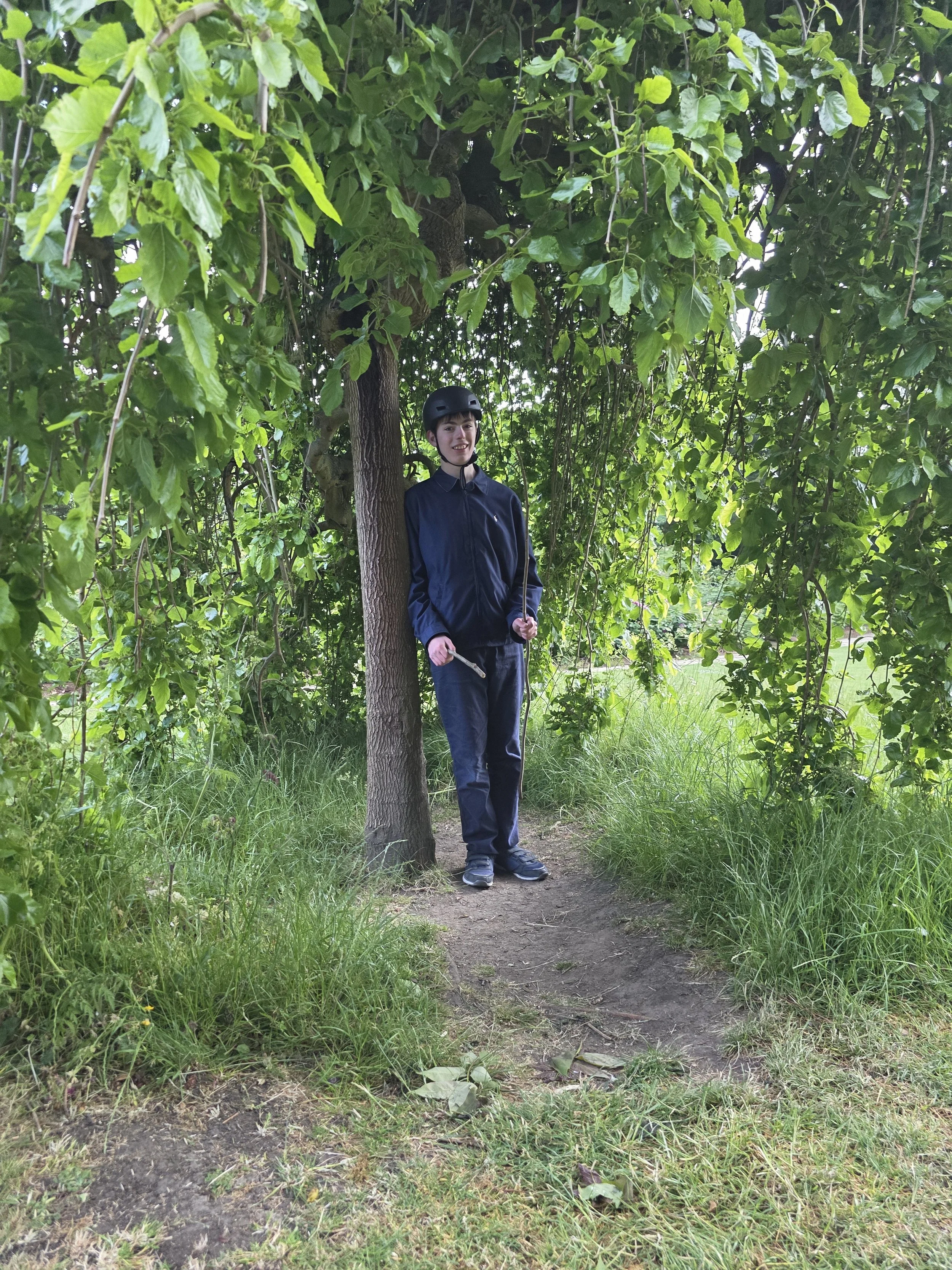 A young person in a black helmet and navy jacket holding a stick, standing next to a tree in a green, leafy outdoor area.