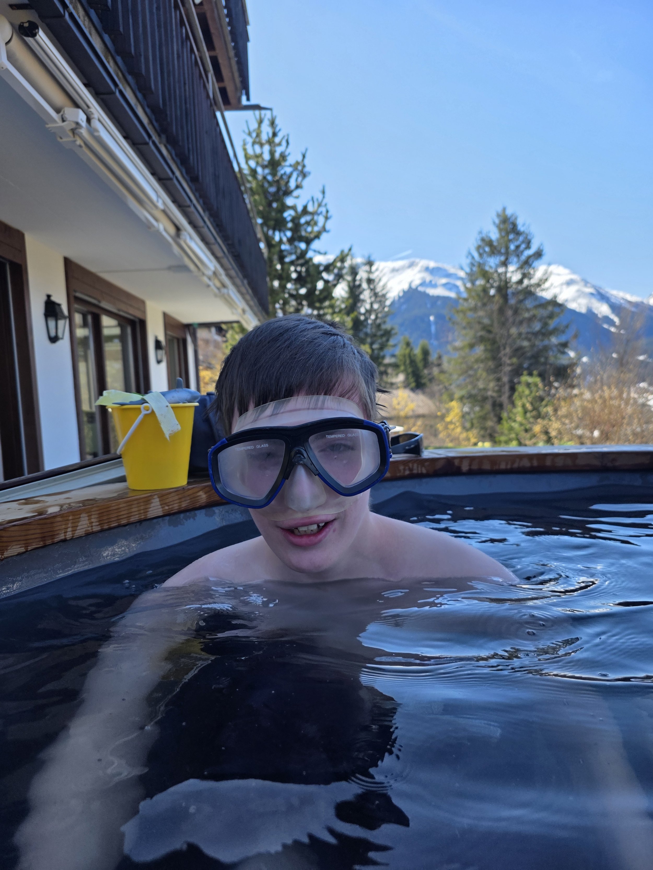 A young boy wearing swimming goggles in an outdoor hot tub with nature and snow-capped mountains in the background.