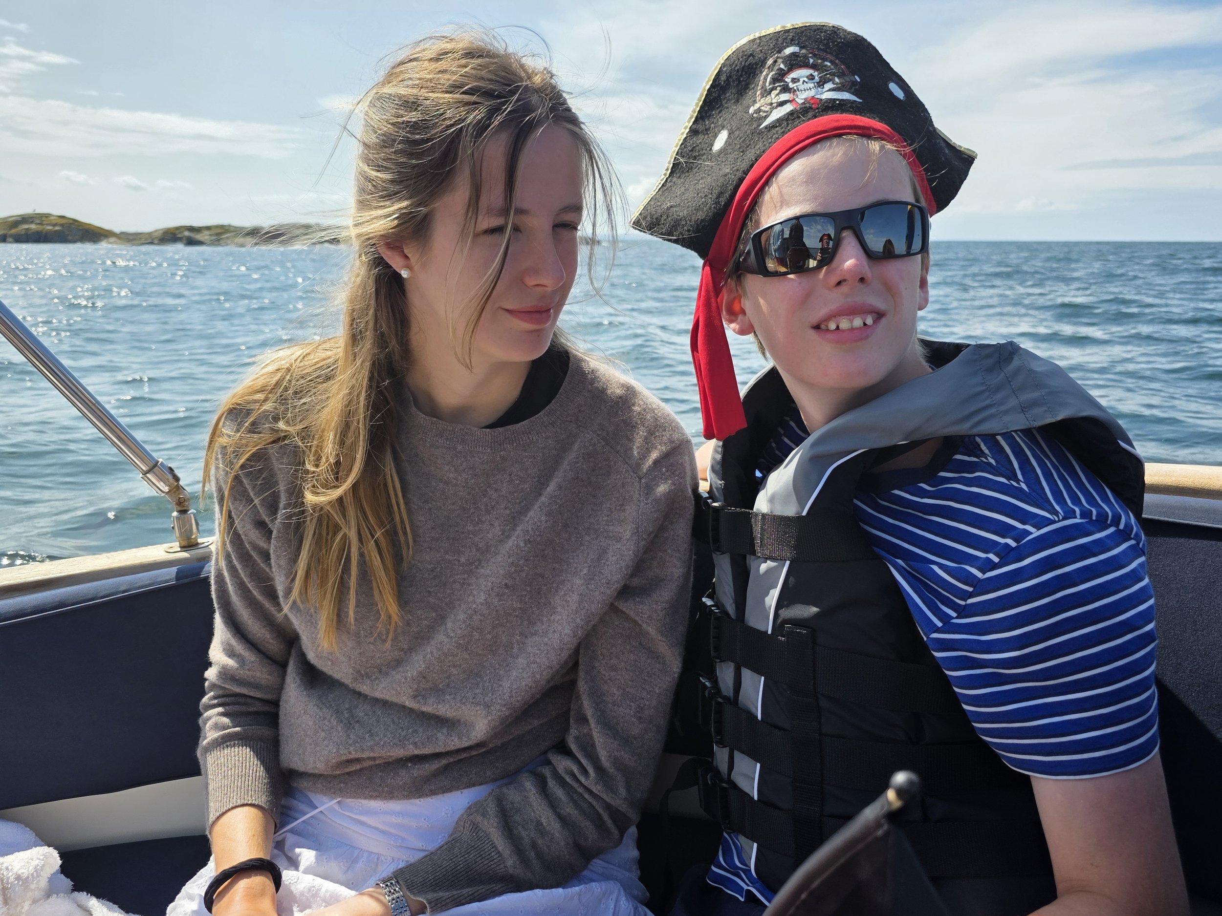 A woman with long blonde hair and a young boy wearing sunglasses and a pirate hat are sitting on a boat, with the ocean and sky in the background.