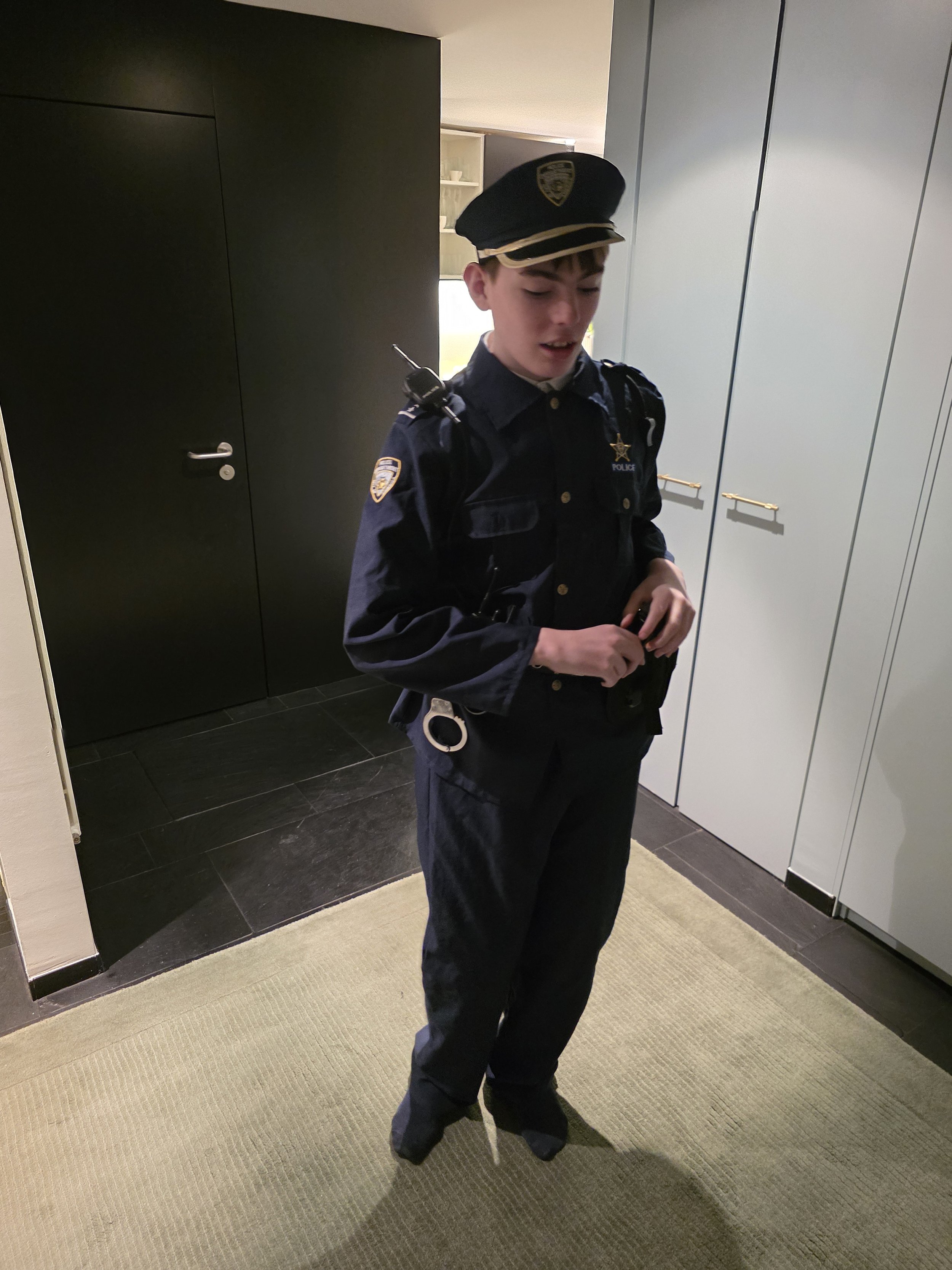 A young man dressed as a police officer stands in a room with gray and beige decor. He is wearing a navy blue police uniform with a badge, a cap with a police emblem, and has a radio on his shoulder. He is looking downward with his eyes closed and his hands clasped.