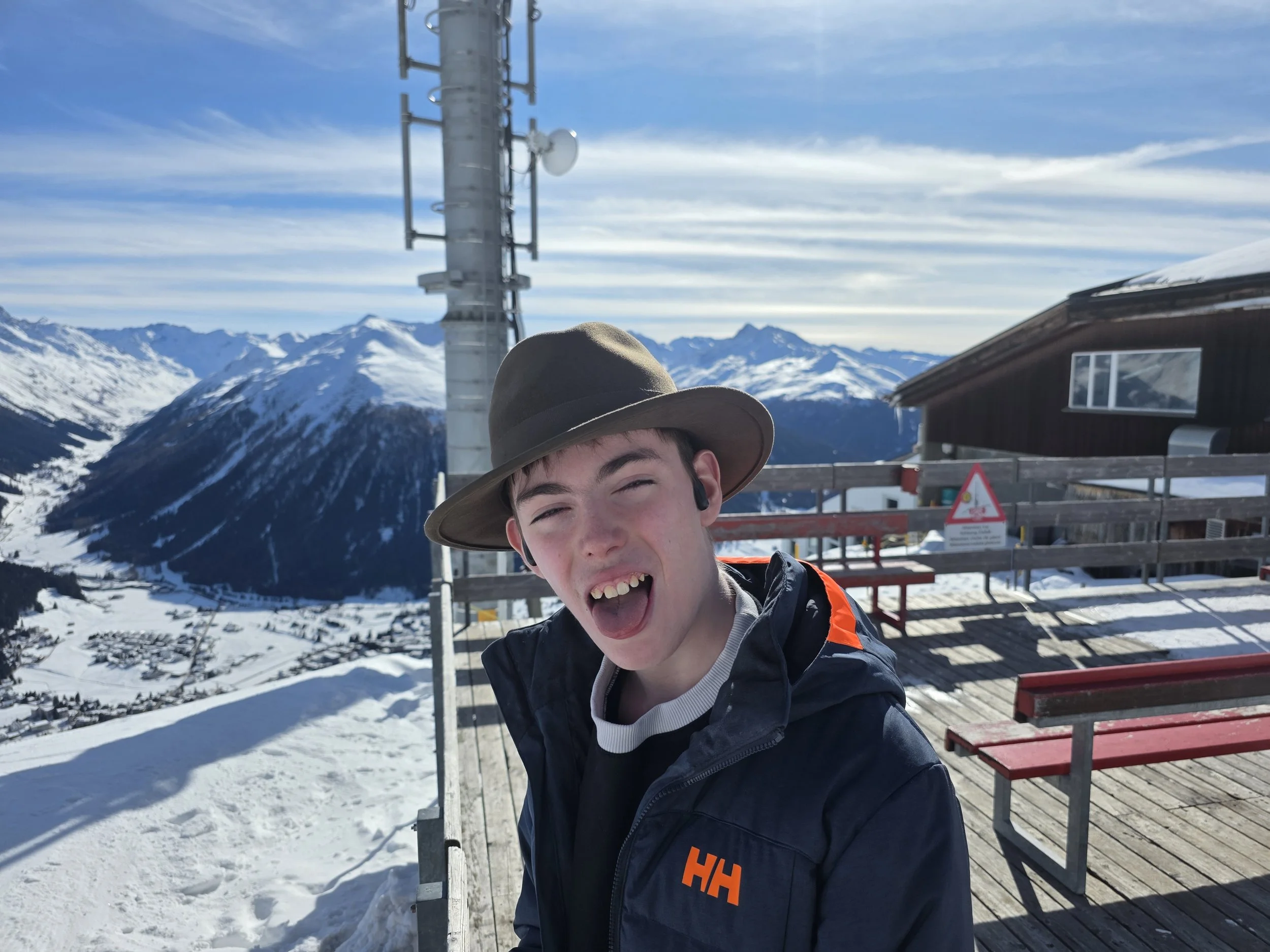 A young man wearing a wide-brimmed hat and a jacket, smiling and taking a selfie in a snowy mountain landscape during daytime. There is a wooden building, red benches, a metal structure with communication equipment, snow-covered mountains in the background, and a partly cloudy sky.
