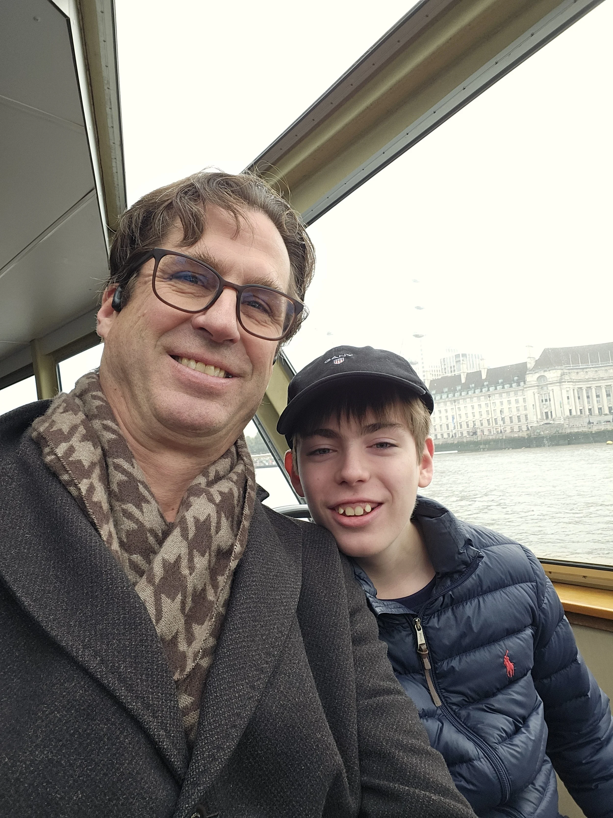 Two males sitting inside a boat near water, with city buildings visible in the background. One is older, wearing glasses and a patterned scarf, smiling at the camera. The other appears younger, wearing a dark cap and a puffy jacket, also smiling.