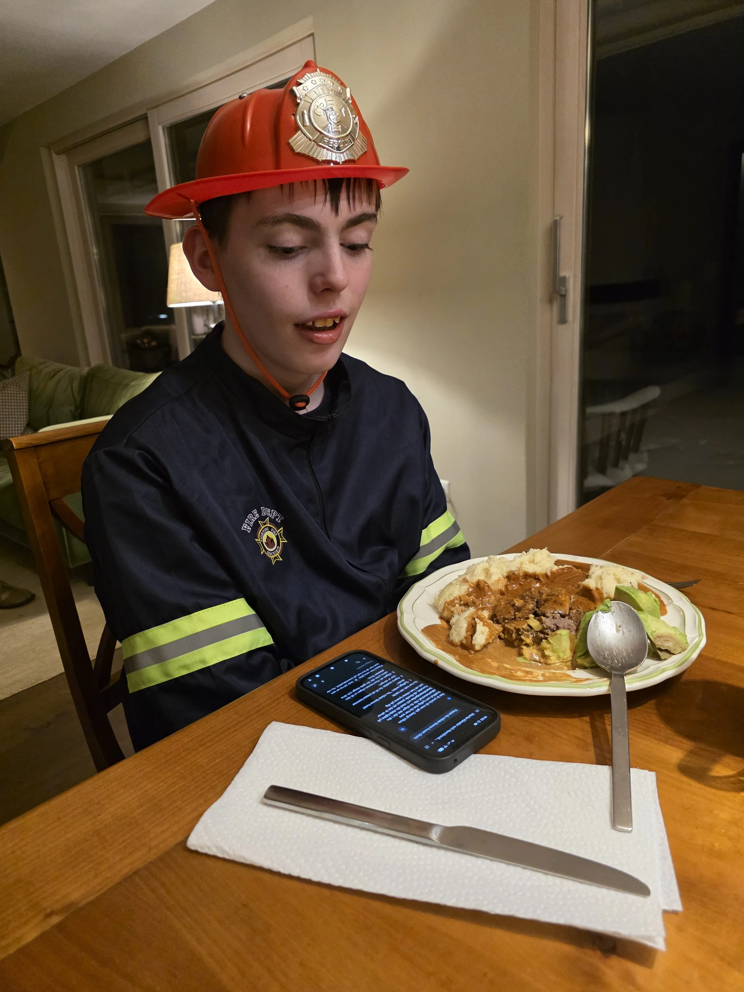A young man wearing a firefighter uniform and an orange firefighter helmet with a badge, sitting at a dining table with a plate of food in front of him, including mashed potatoes, gravy, and vegetables.