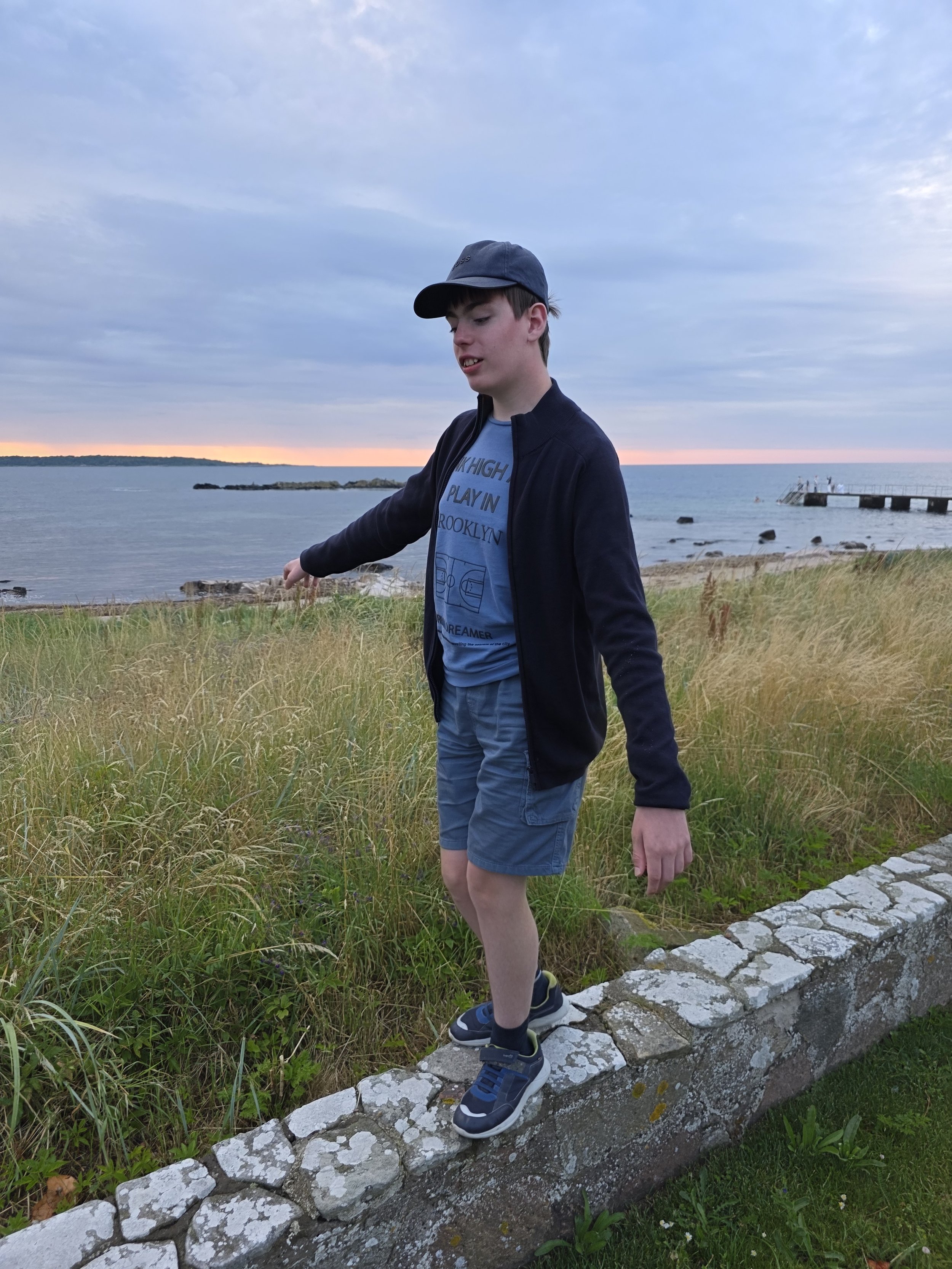 A teenage boy standing on a stone wall near the water with grass and a pier in the background during sunset.