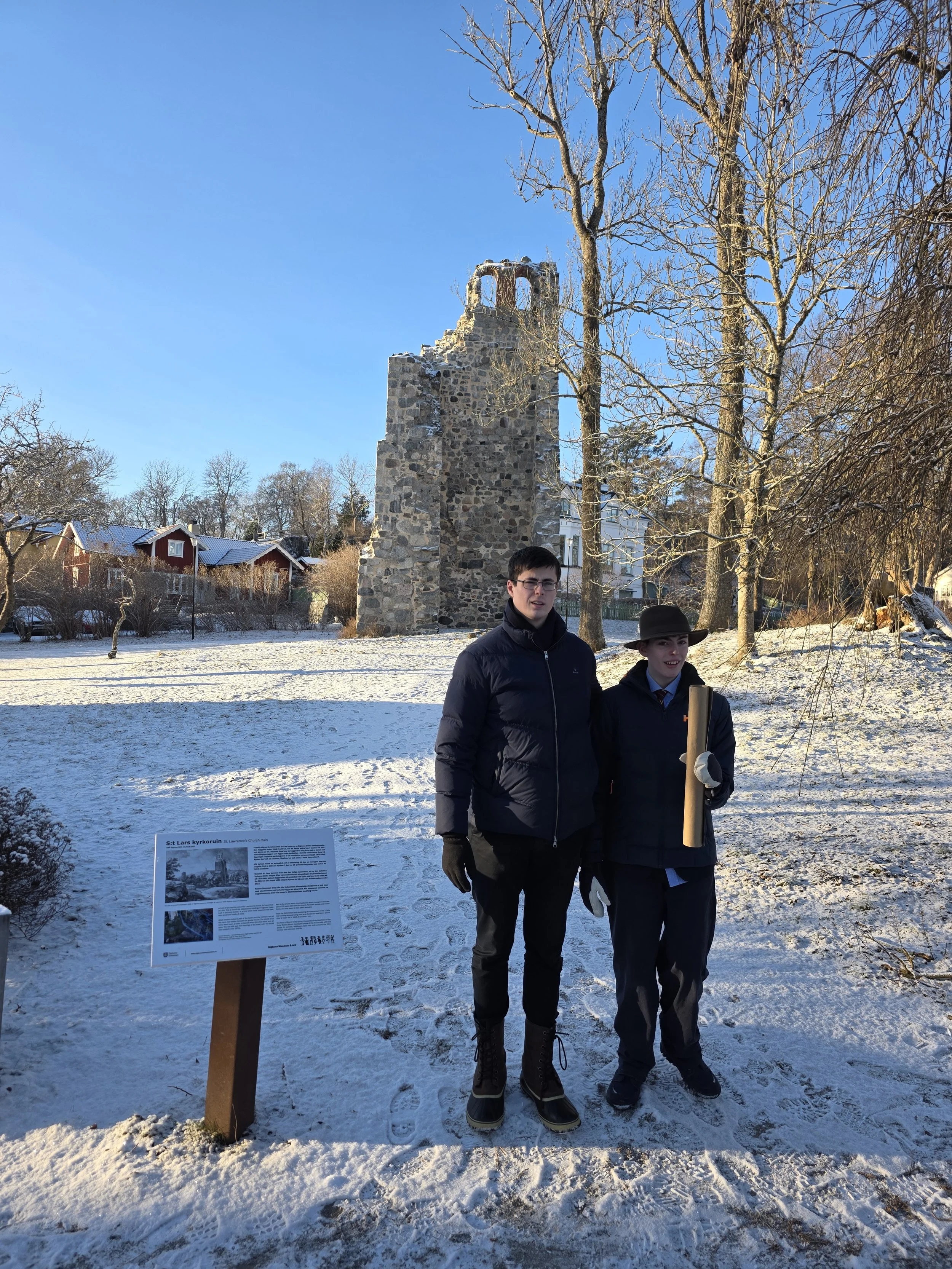 Two young men standing outdoors in a snowy area with bare trees. One holds a rolled-up document. Behind them is a stone church ruin with an informational sign in front of it.