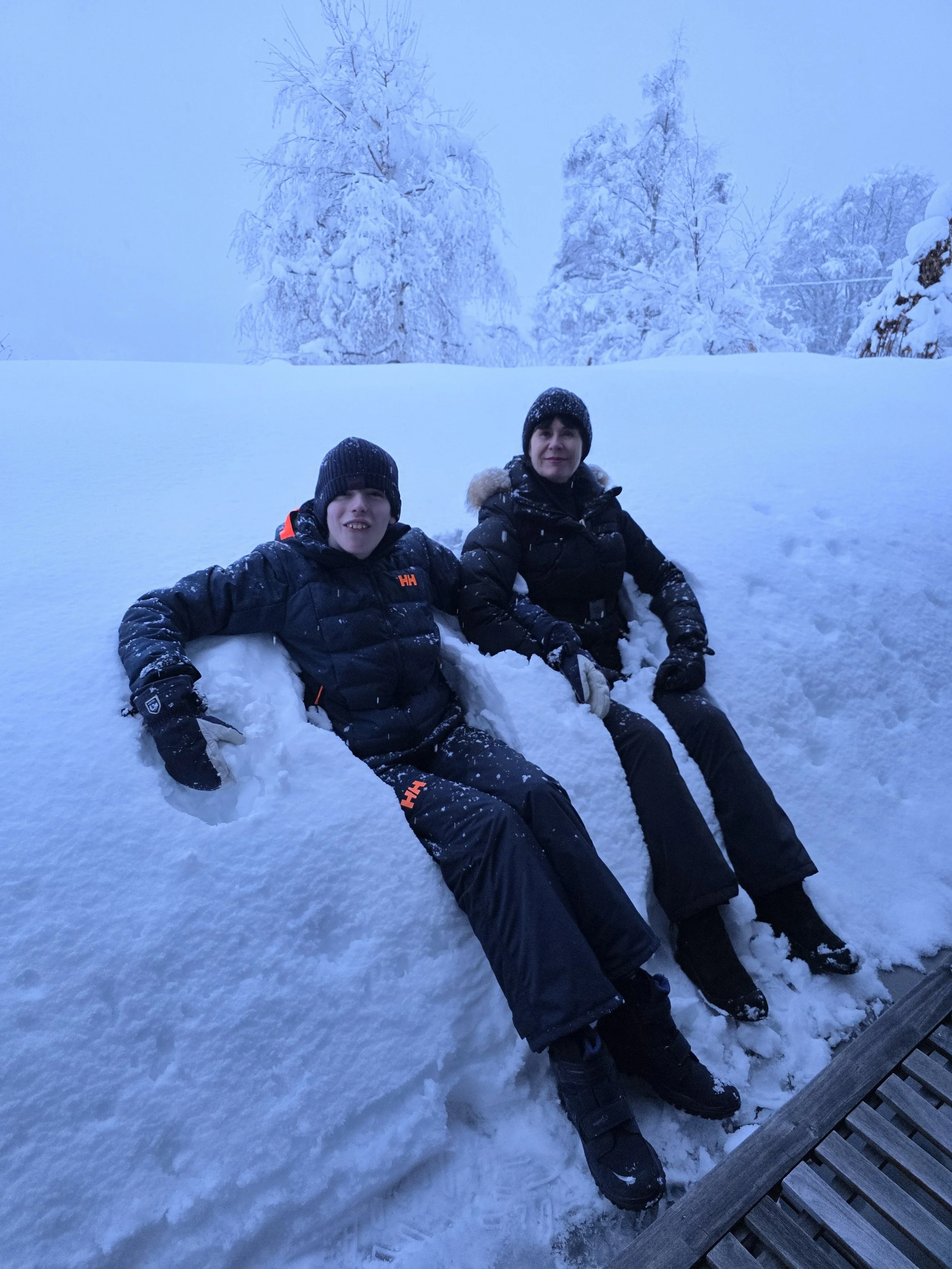 Two people in winter clothing sitting on snow in a snowy outdoor setting, with snow-covered trees in the background.
