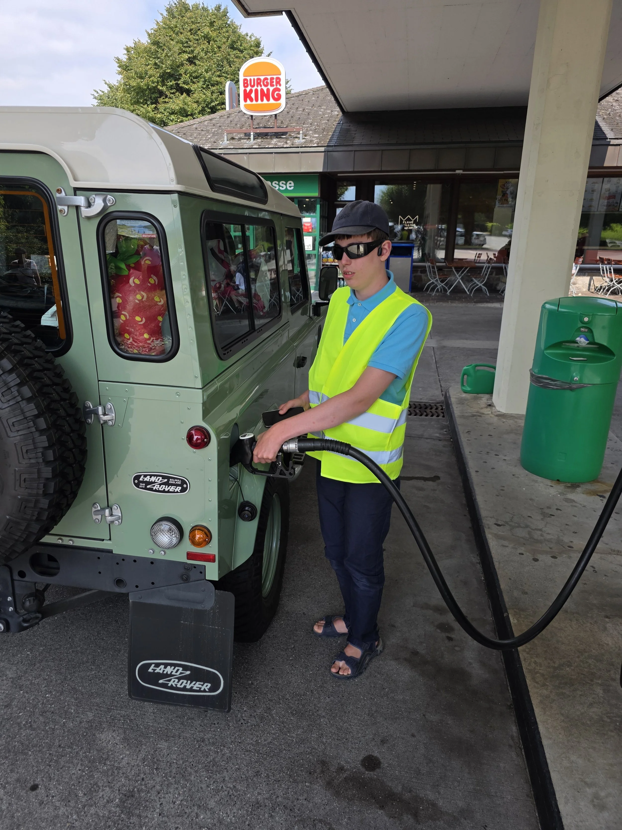 A young man wearing a yellow safety vest, sunglasses, a dark cap, a blue polo shirt, and sandals is filling up a green Land Rover vehicle with fuel at a gas station. The background shows a Burger King sign, a building with potted plants and outdoor seating, and a tree.