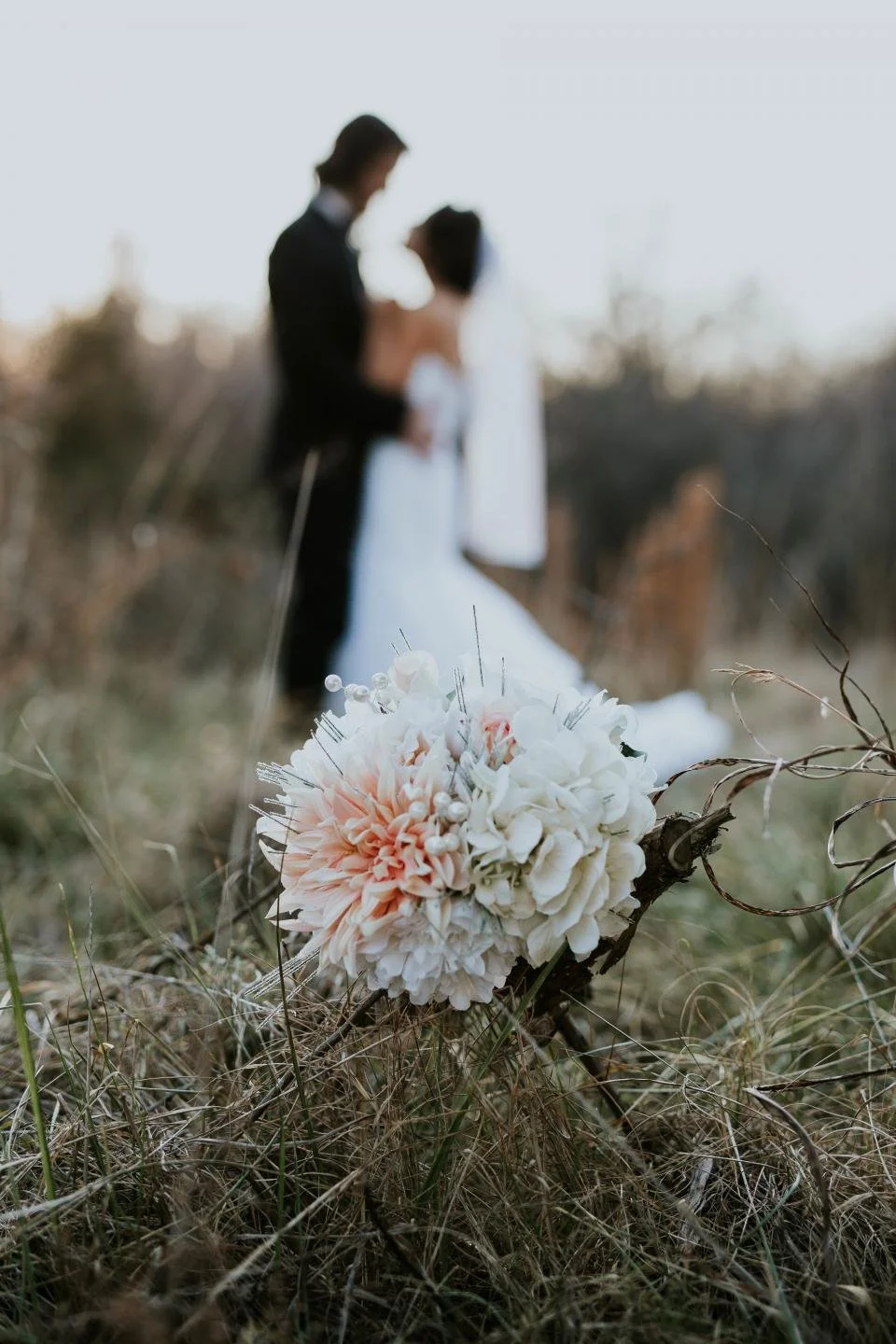 Close-up of a wedding bouquet with pink and white flowers on the ground, with a blurred bride and groom in the background, outdoors in a field. Imagine your own wedding at Grand Shores wedding venue.