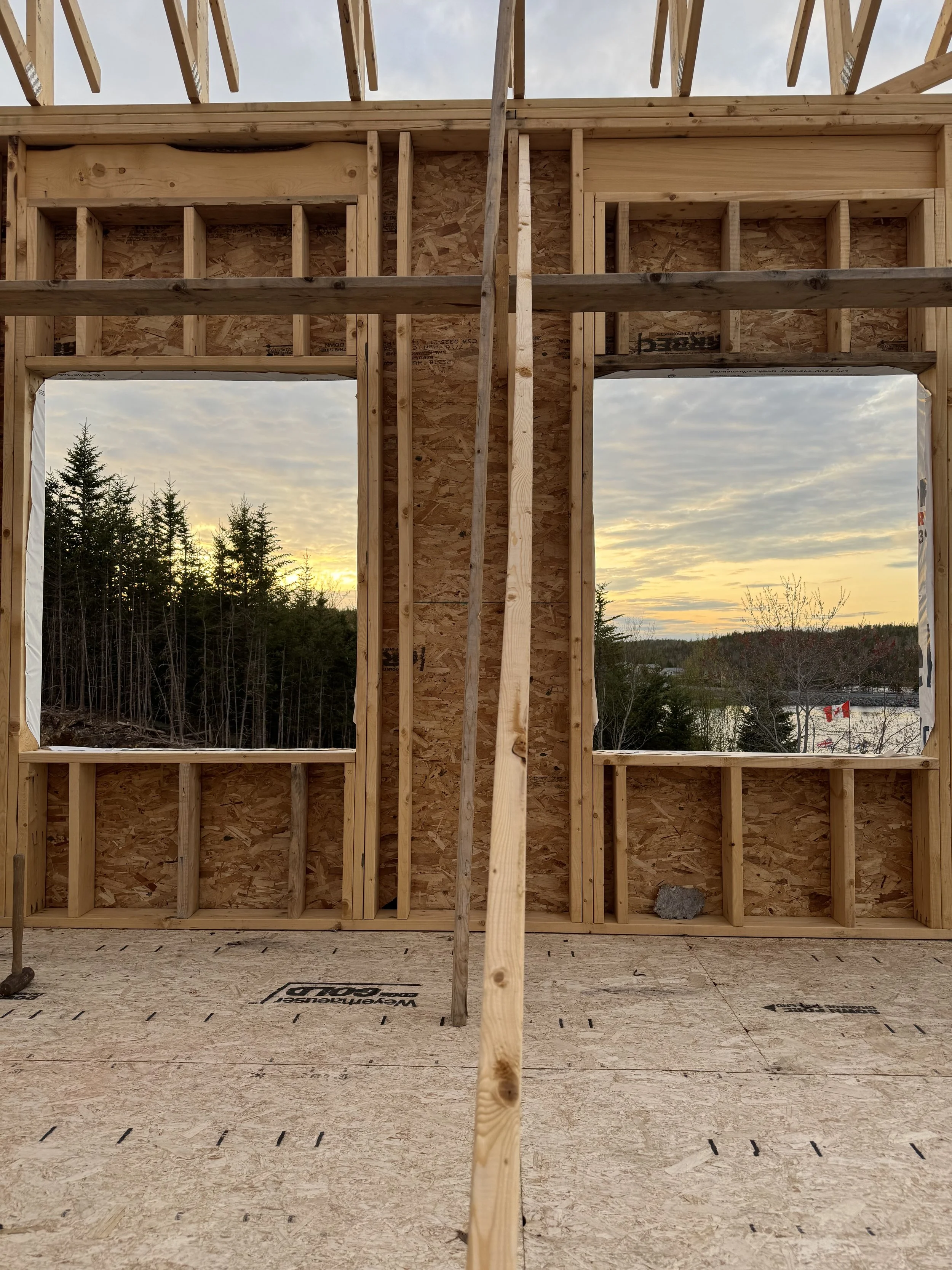 View of the wooden framing of Grand Shores event venue under construction with two windows and a doorway, overlooking trees and a sunset sky.