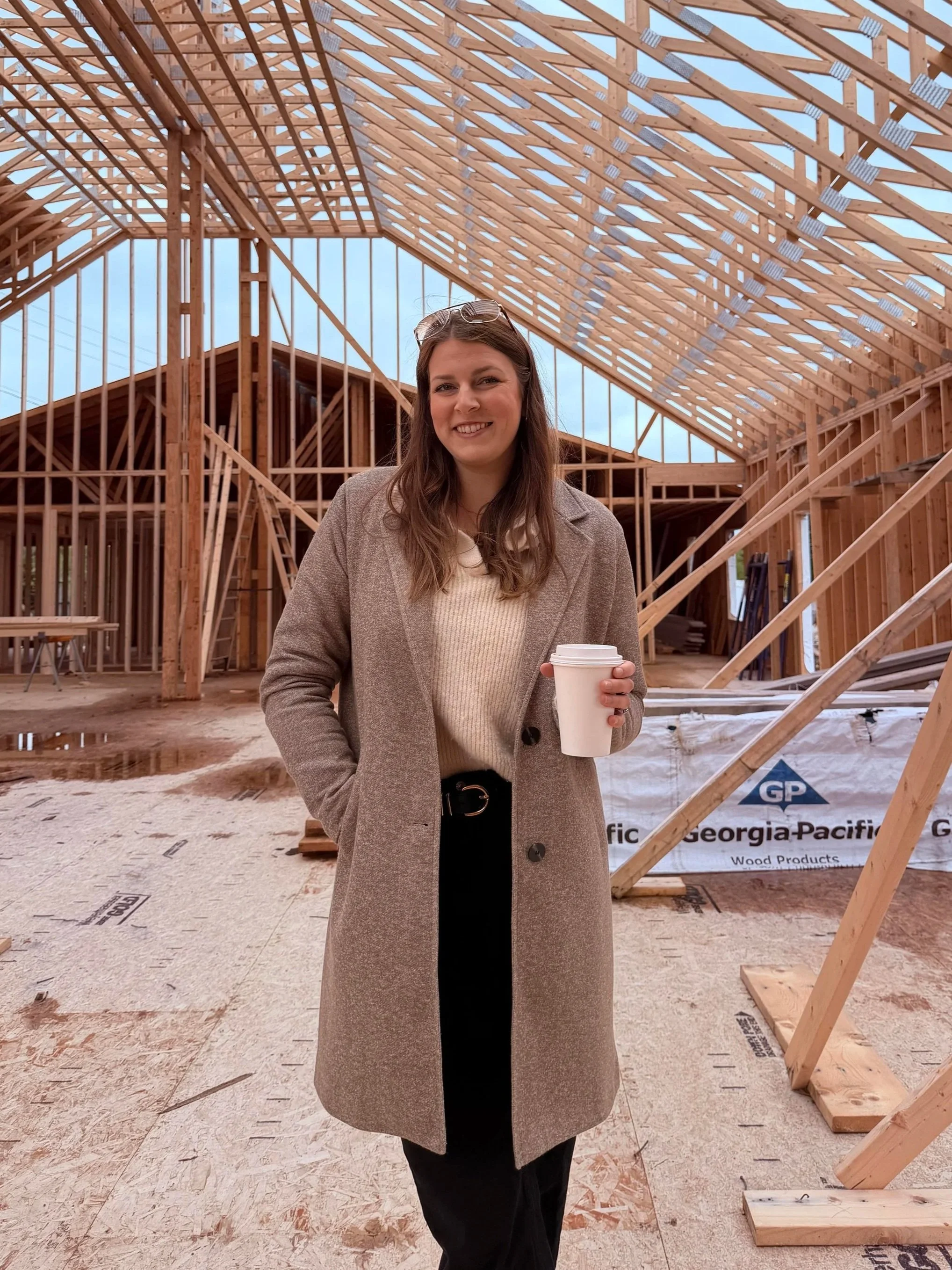 A woman, Colby Baker, co-owner of the Grand Shores wedding venue in Nova Scotia, stands inside a wooden frame construction site, holding a coffee cup and smiling, wearing a gray coat and glasses.