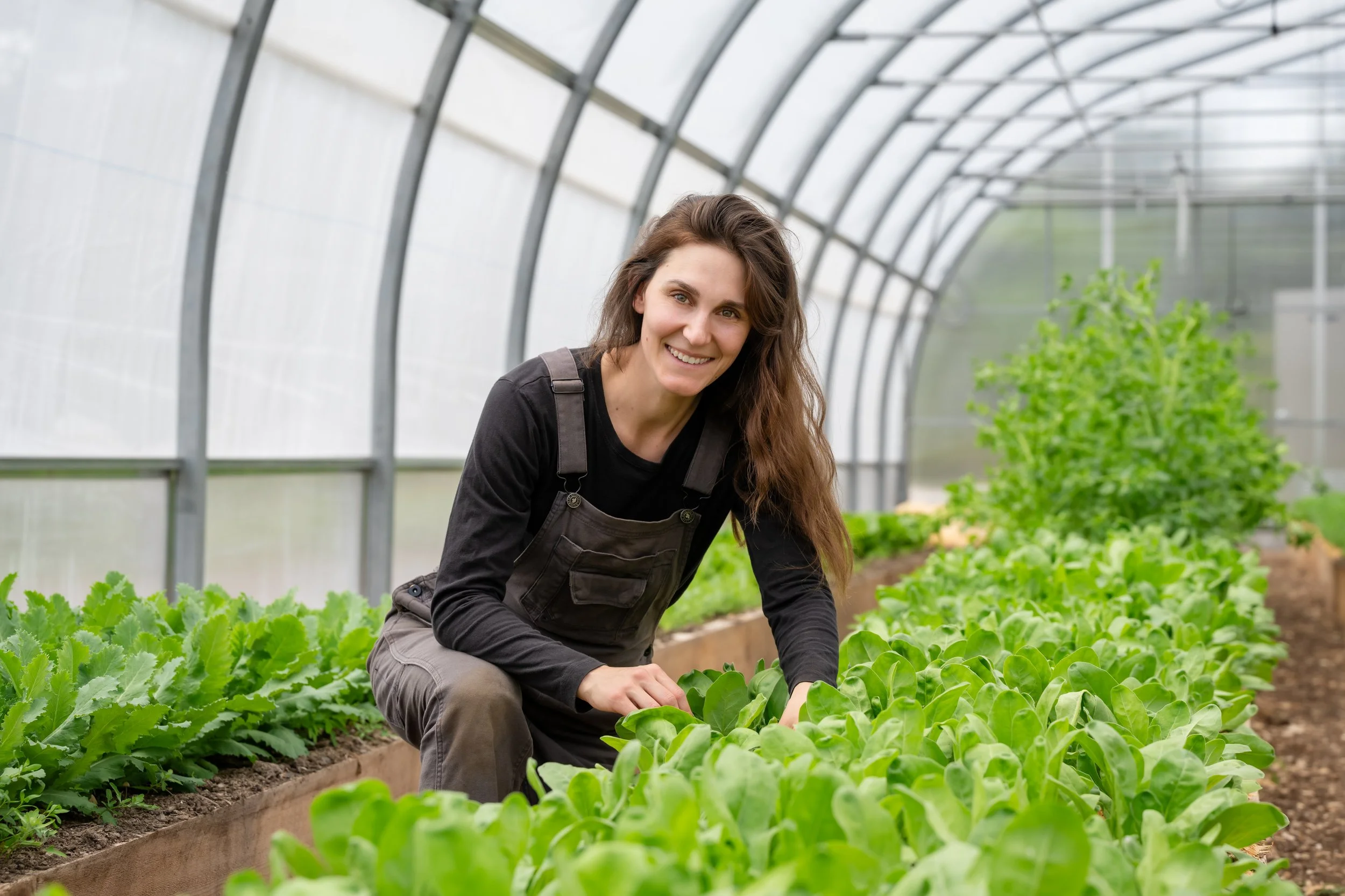 A woman, Erika Clarke, co-owner of the Grand Shores wedding venue in Nova Scotia, with long brown hair, smiling while farming inside a greenhouse with green leafy plants.