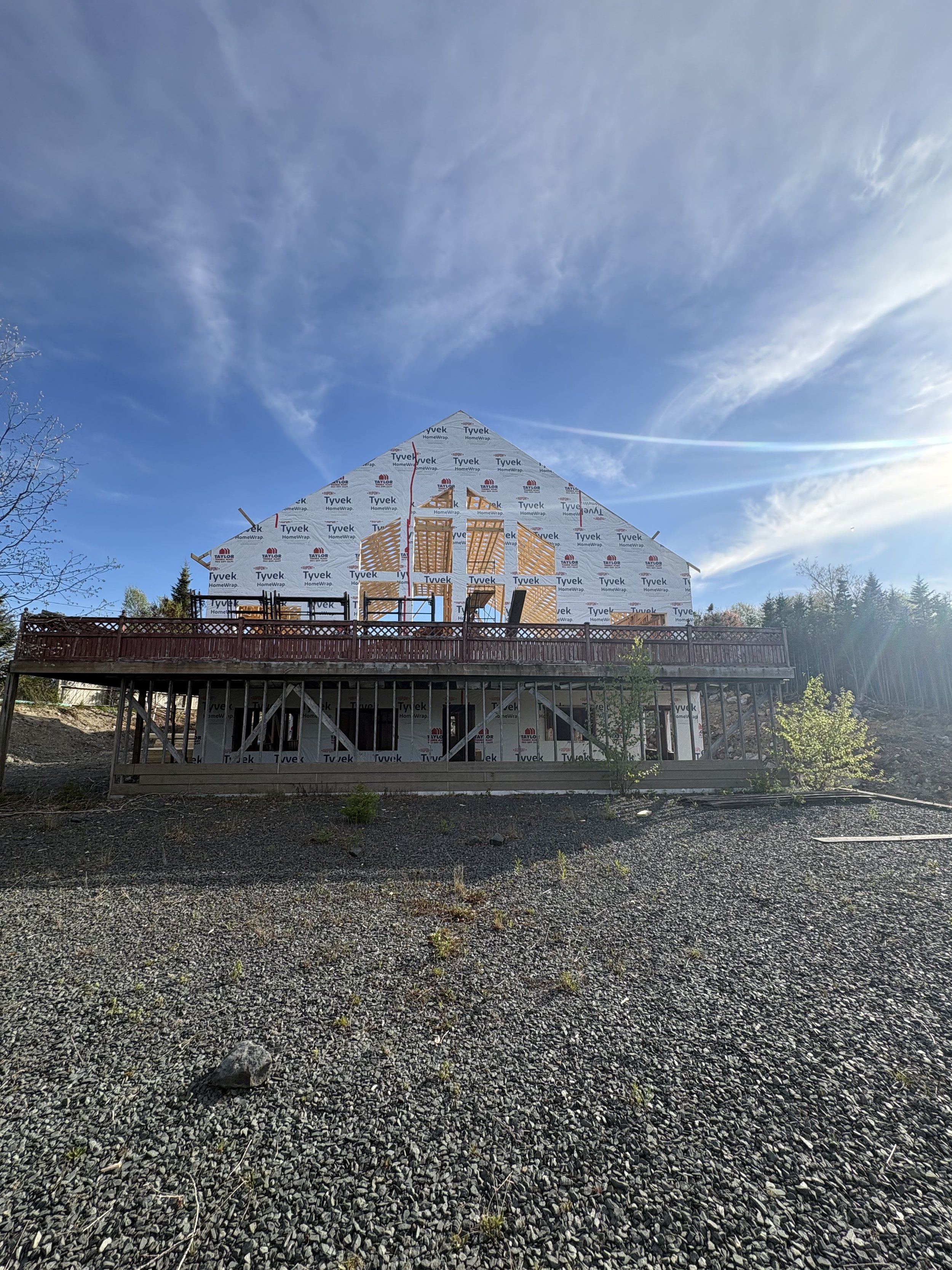 Wedding venue, Grand Shores, under construction with exposed wooden framing and Tyvek wrap, surrounded by a fence, with gravel ground and trees in the background under a partly cloudy sky.