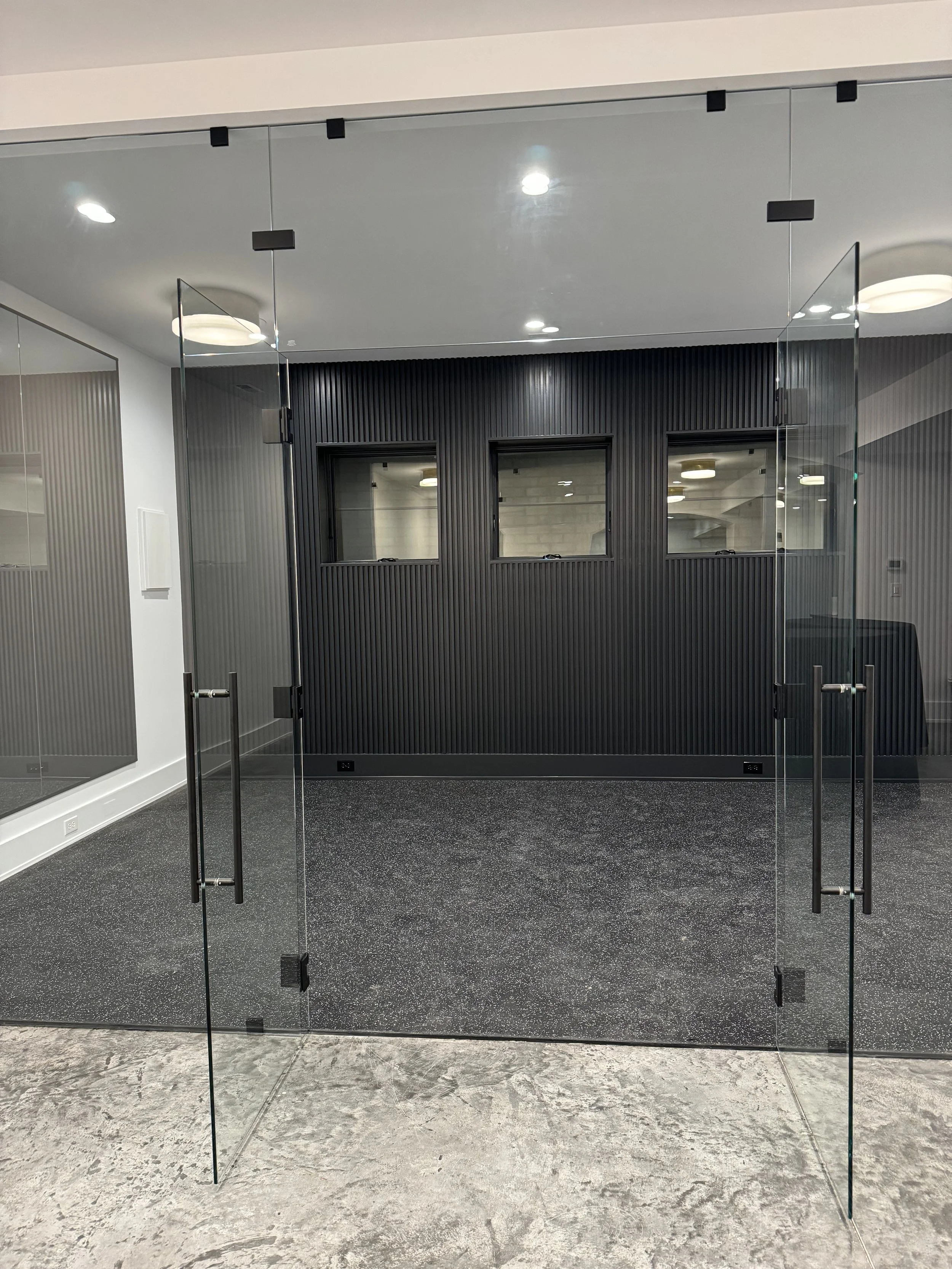 Glass door opens into a modern office or lobby with black textured wall and three windows above, two closed, one open, with a dark speckled floor and white walls.