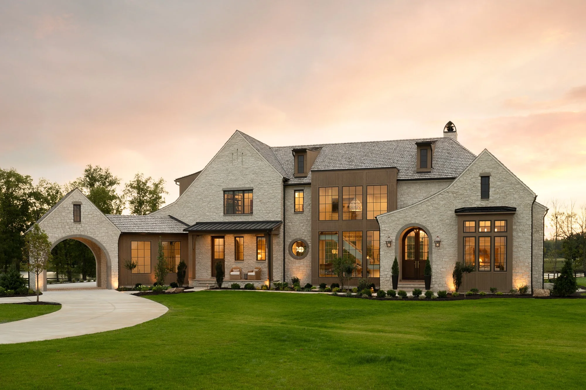 Large modern house with stone and wood exterior, large windows, and a manicured lawn at sunset.