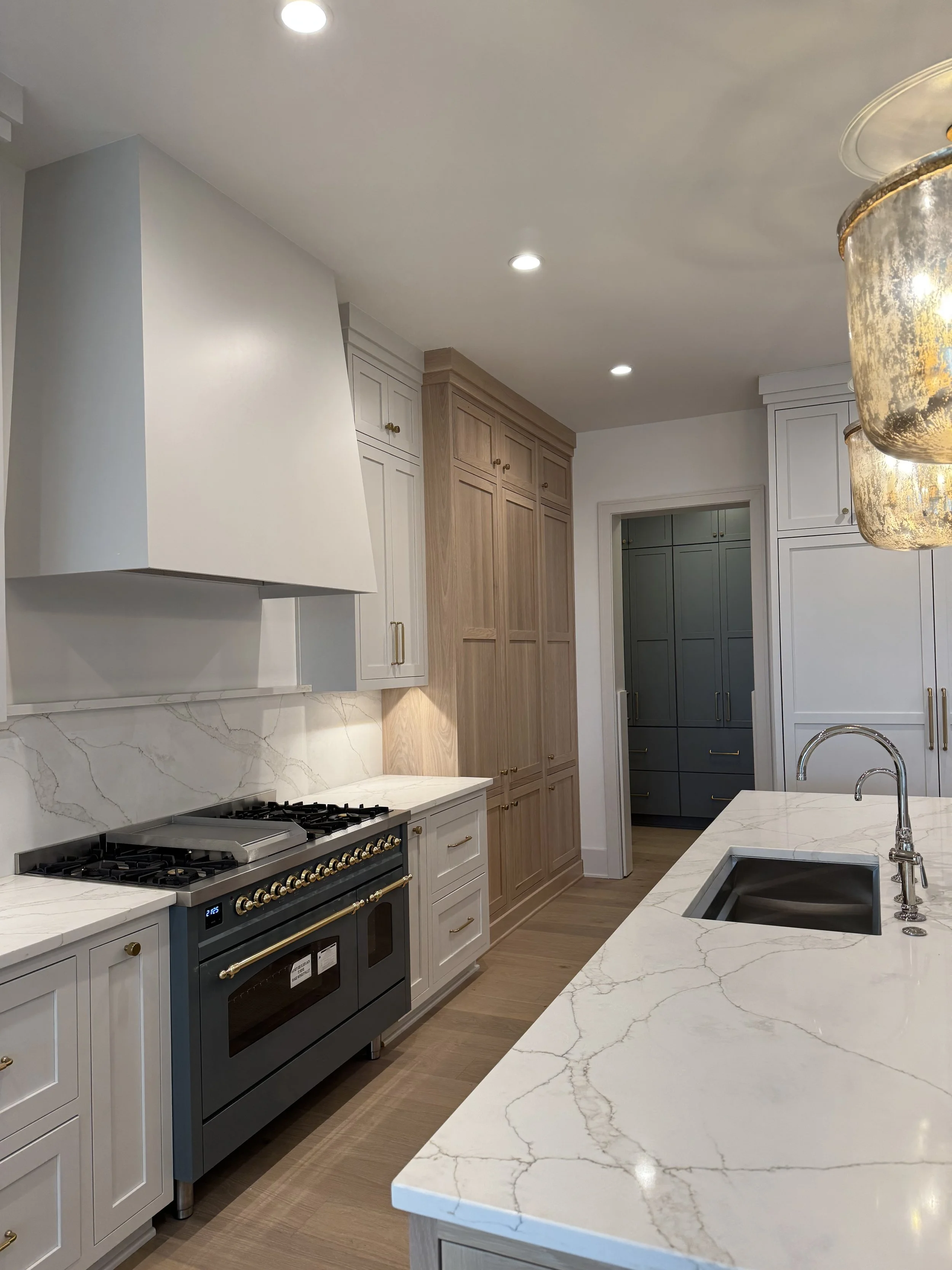 A modern kitchen with white and wood cabinets, marble countertops, a black stove, and pendant lighting.