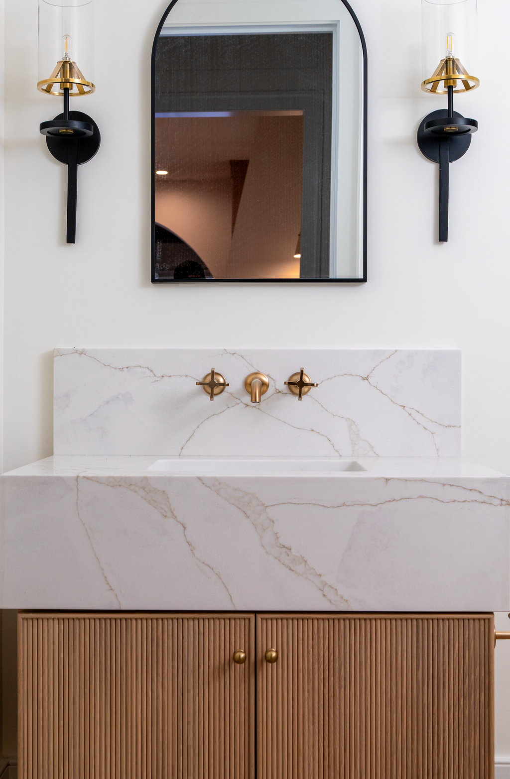 Modern bathroom vanity with a white marble sink and gold fixtures, a rectangular mirror, and black and gold wall sconces.