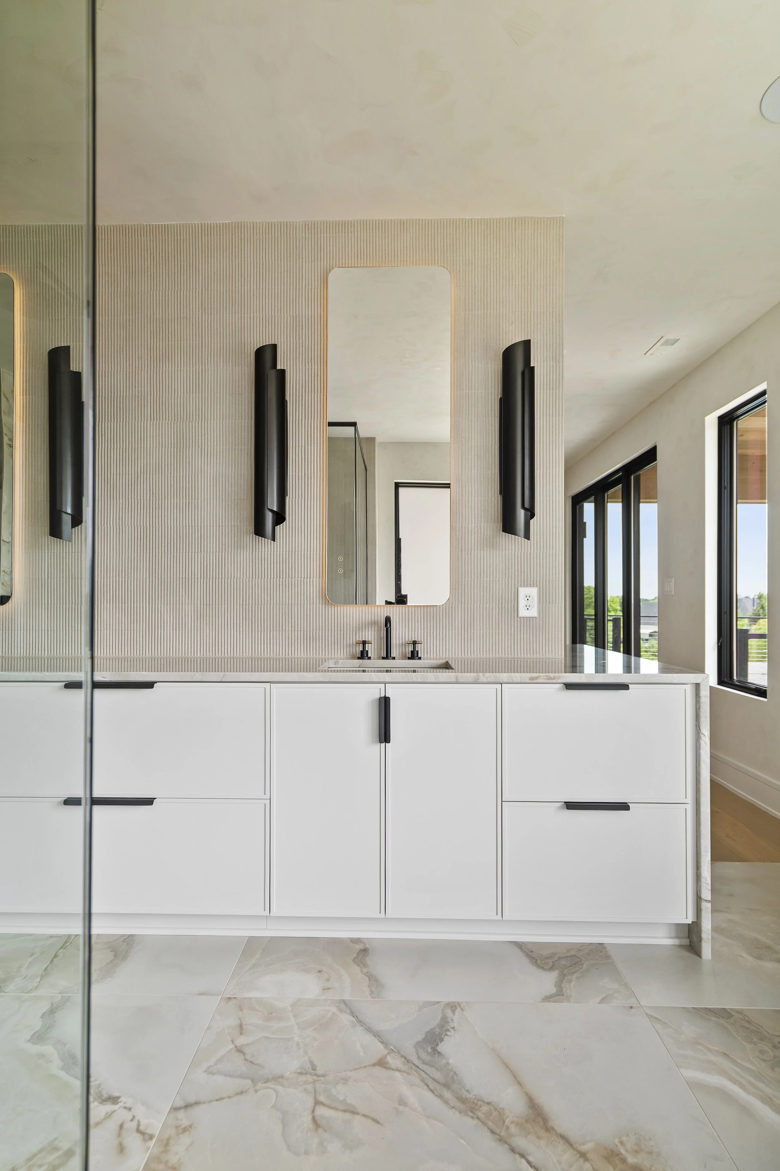 Modern bathroom with white vanity, black fixtures, a large mirror, and beige textured wall tiles. There are black wall sconces on each side of the mirror, and natural light from sliding glass doors.