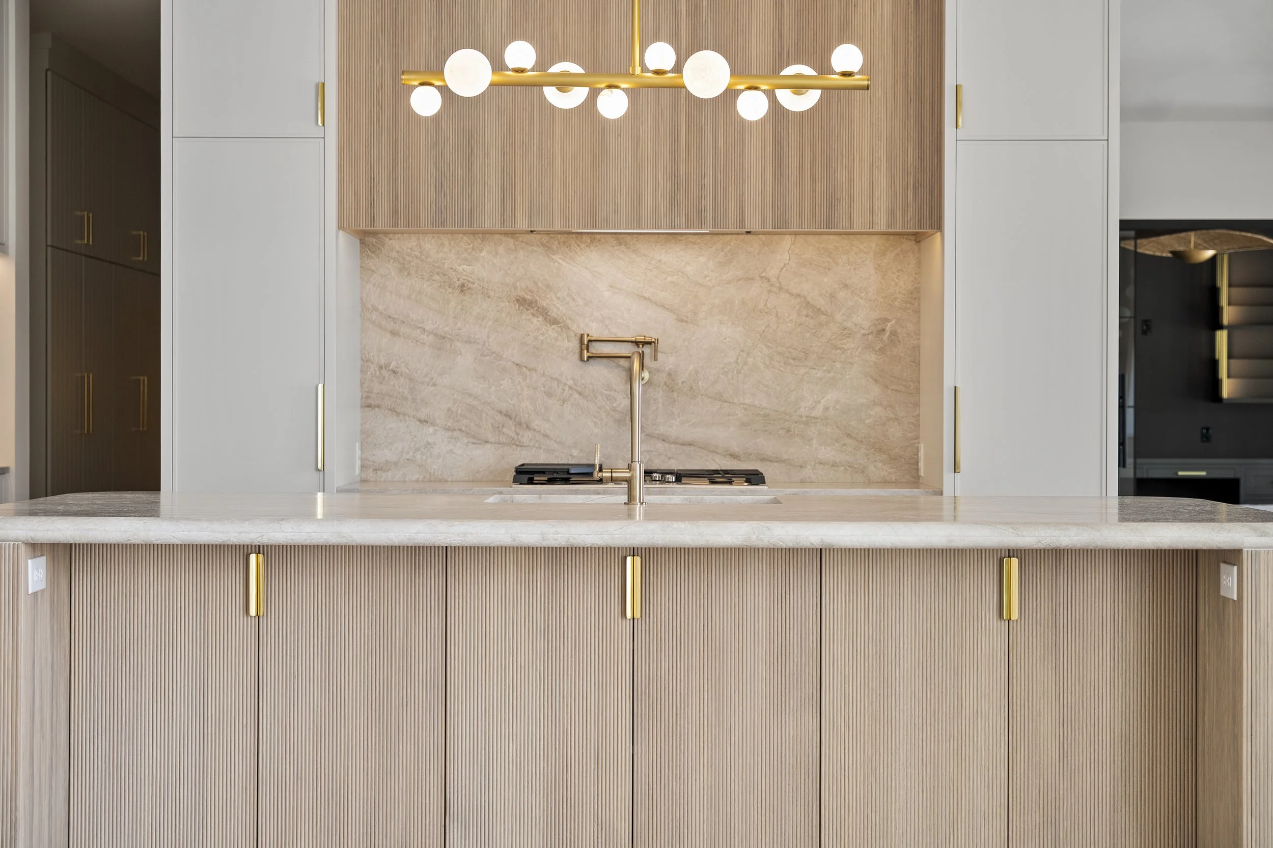 Modern kitchen with a beige marble backsplash, wooden upper cabinets, white lower cabinets with gold handles, and a gold faucet. A gold hanging chandelier with globe lights is above the countertop.