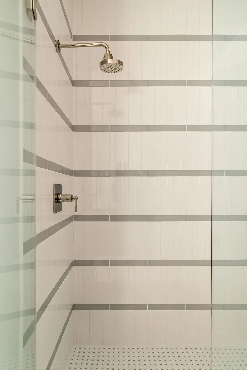 Empty walk-in shower with white tile walls, gray horizontal accent lines, a stainless steel showerhead, and a silver control knob.
