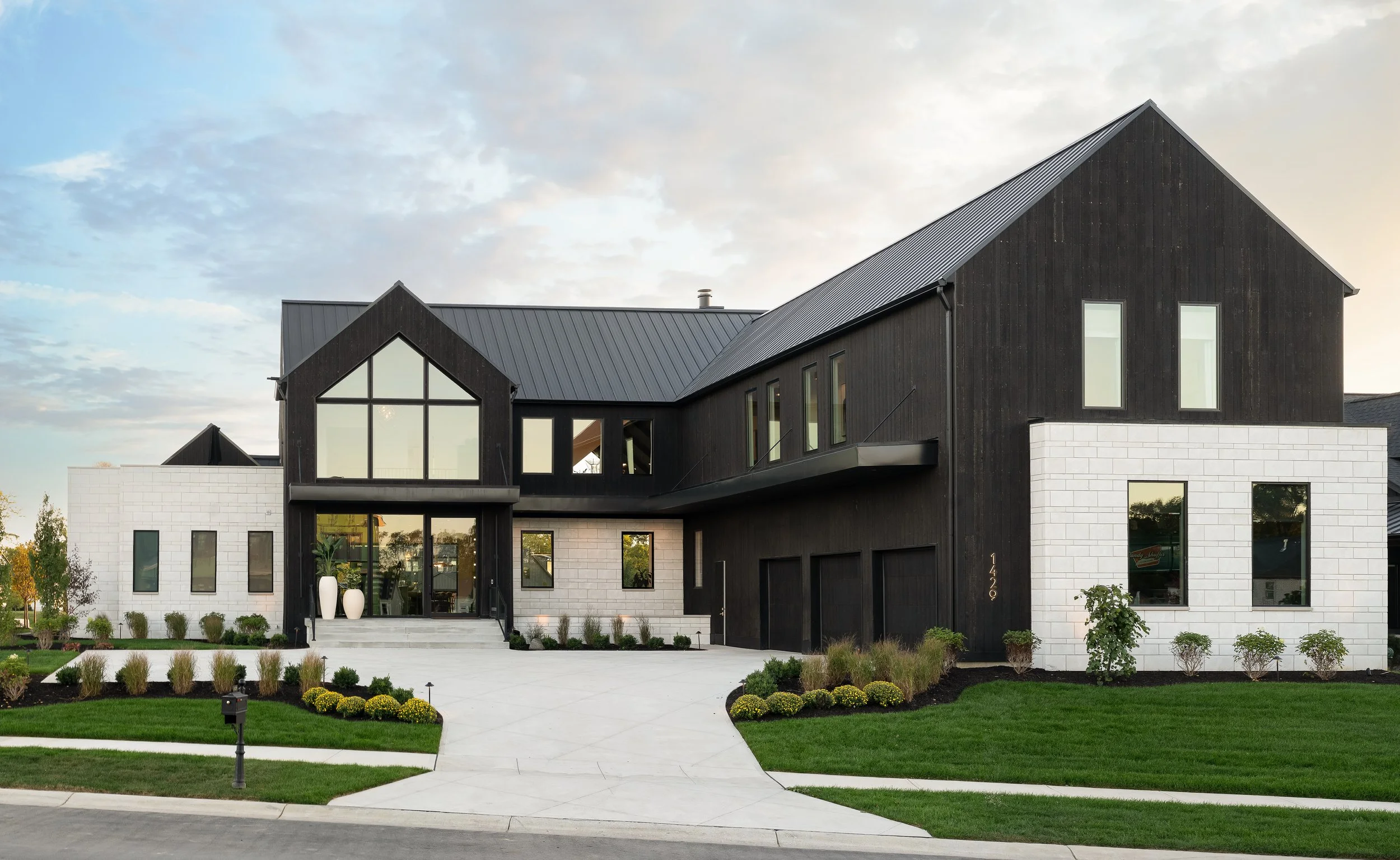Modern two-story house with black wooden exterior, white stone accents, large windows, front porch with stairs, landscaped front yard with grass, small bushes, and a sidewalk, under a partly cloudy sky.