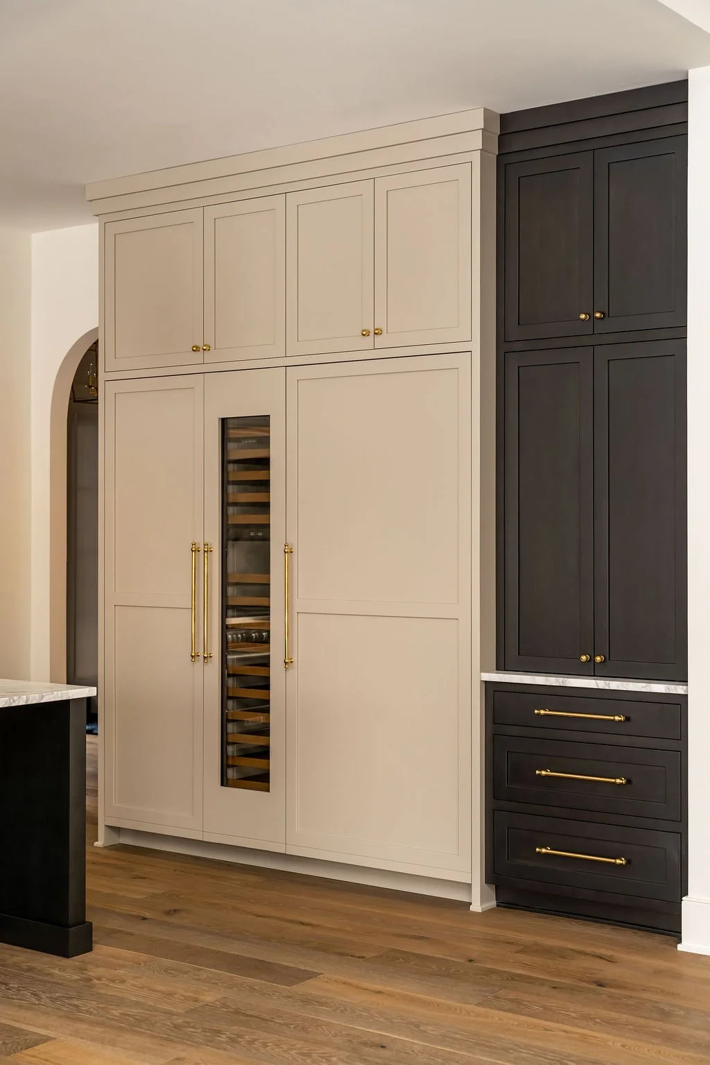 Kitchen cabinetry with beige upper and lower cabinets, black pantry cabinets with gold handles, a built-in wine cooler, and wood flooring.